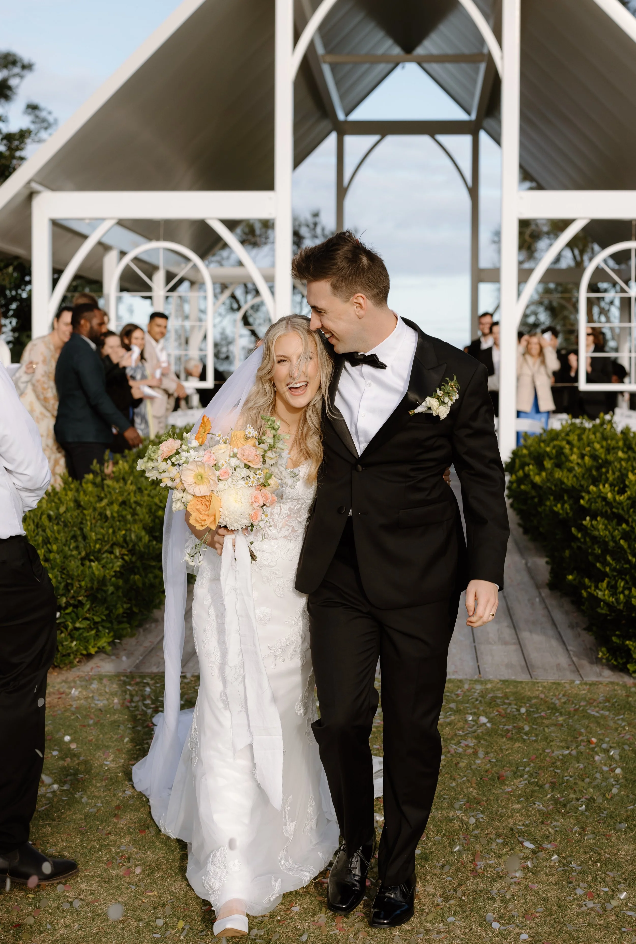 Bride and groom walking arm-in-arm down the aisle at their outdoor wedding, smiling and laughing, with friends and family in the background, under a white wedding arch.