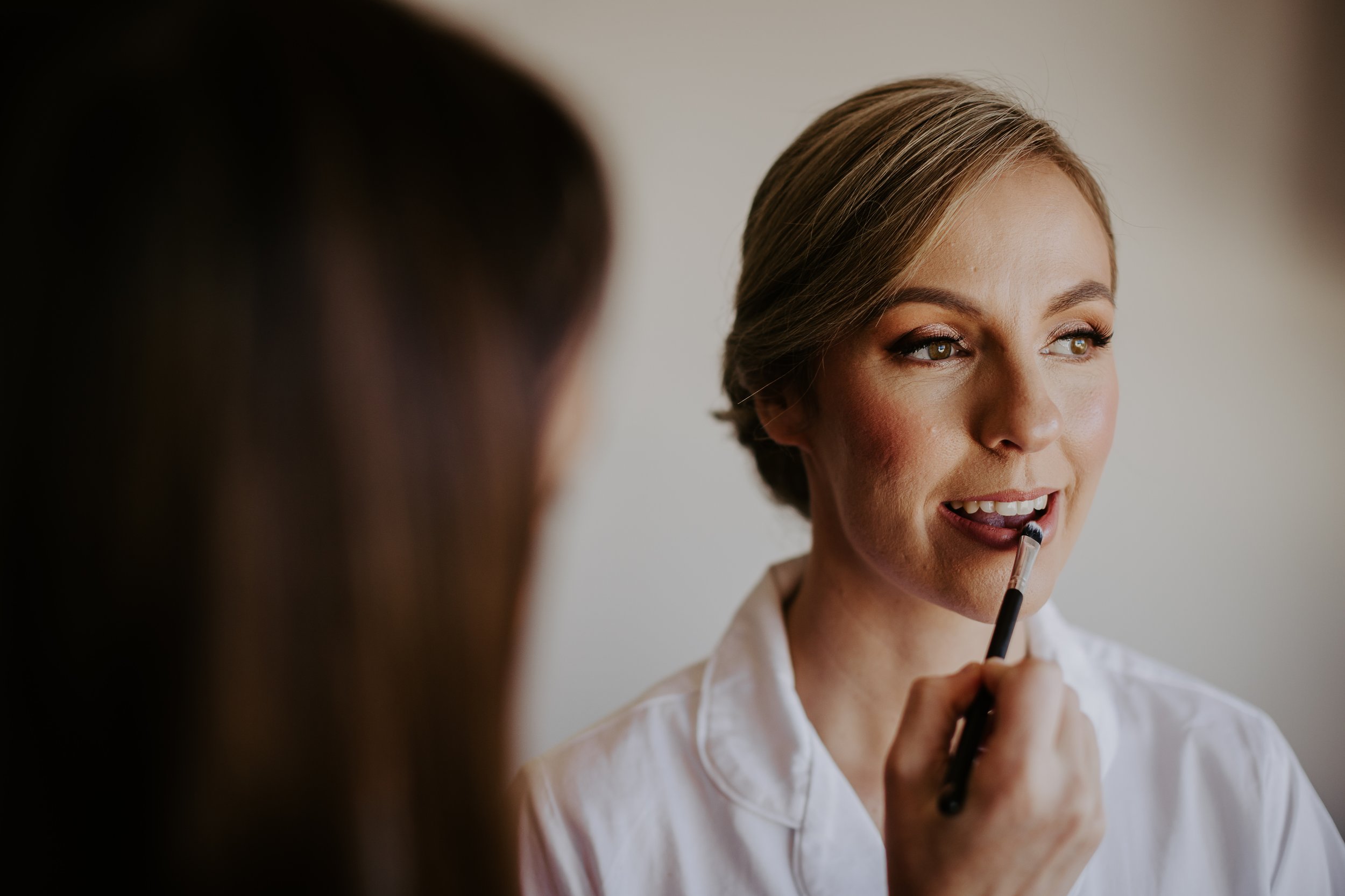 A woman with light brown hair getting makeup applied, holding a makeup brush near her lips, with another person partially visible applying makeup.