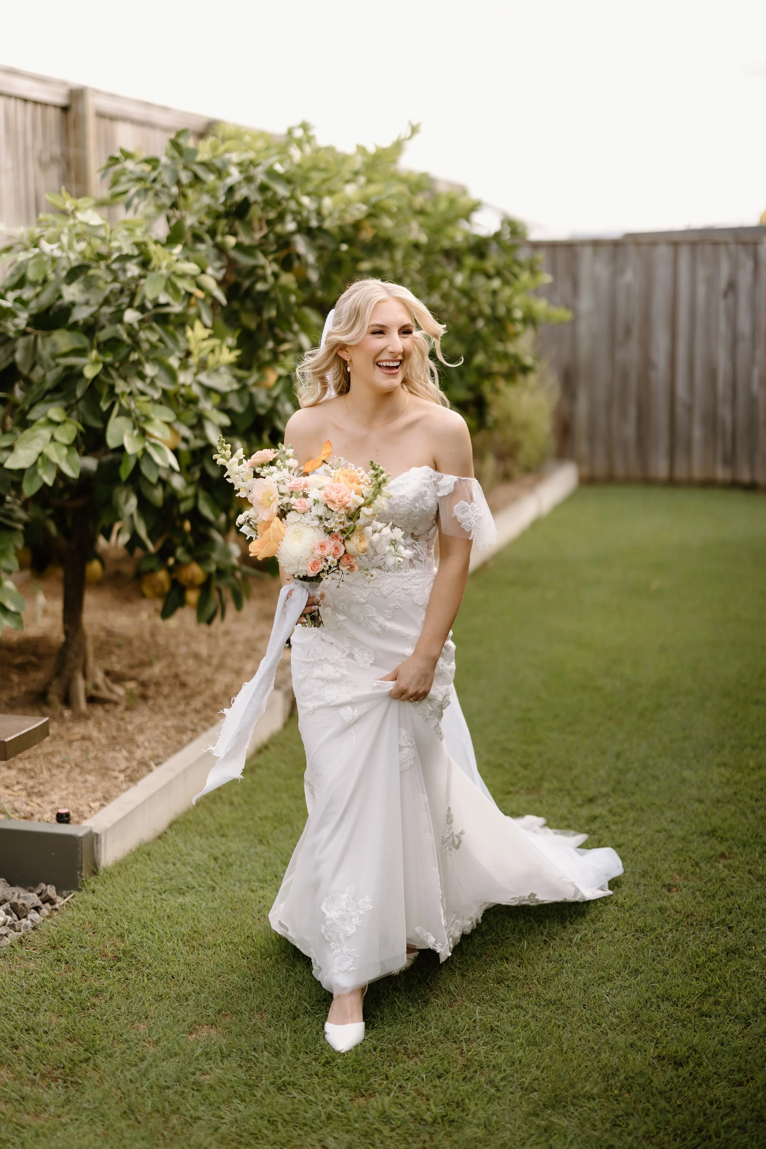 A smiling bride in a white lace wedding dress holding a bouquet of pink, peach, and white flowers in a backyard with green grass, trees, and a wooden fence.