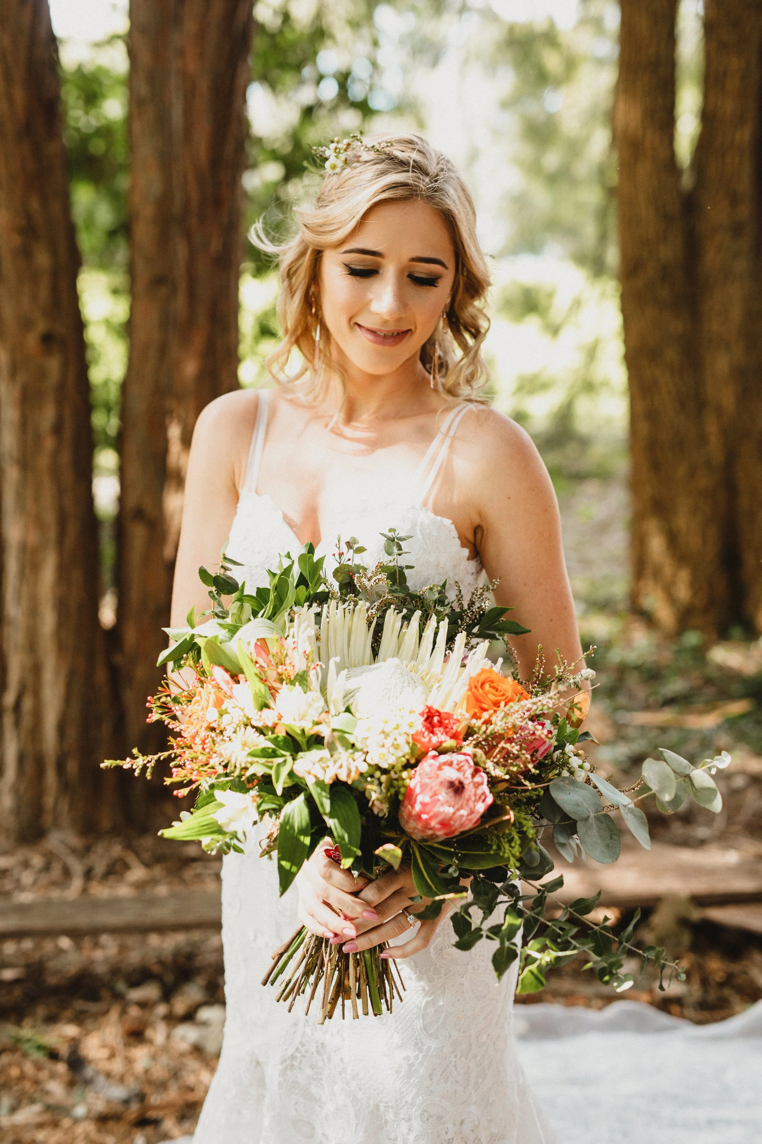 A bride with blonde hair holding a colorful bouquet of flowers standing in a wooded outdoor setting, wearing a white lace wedding dress and smiling softly.