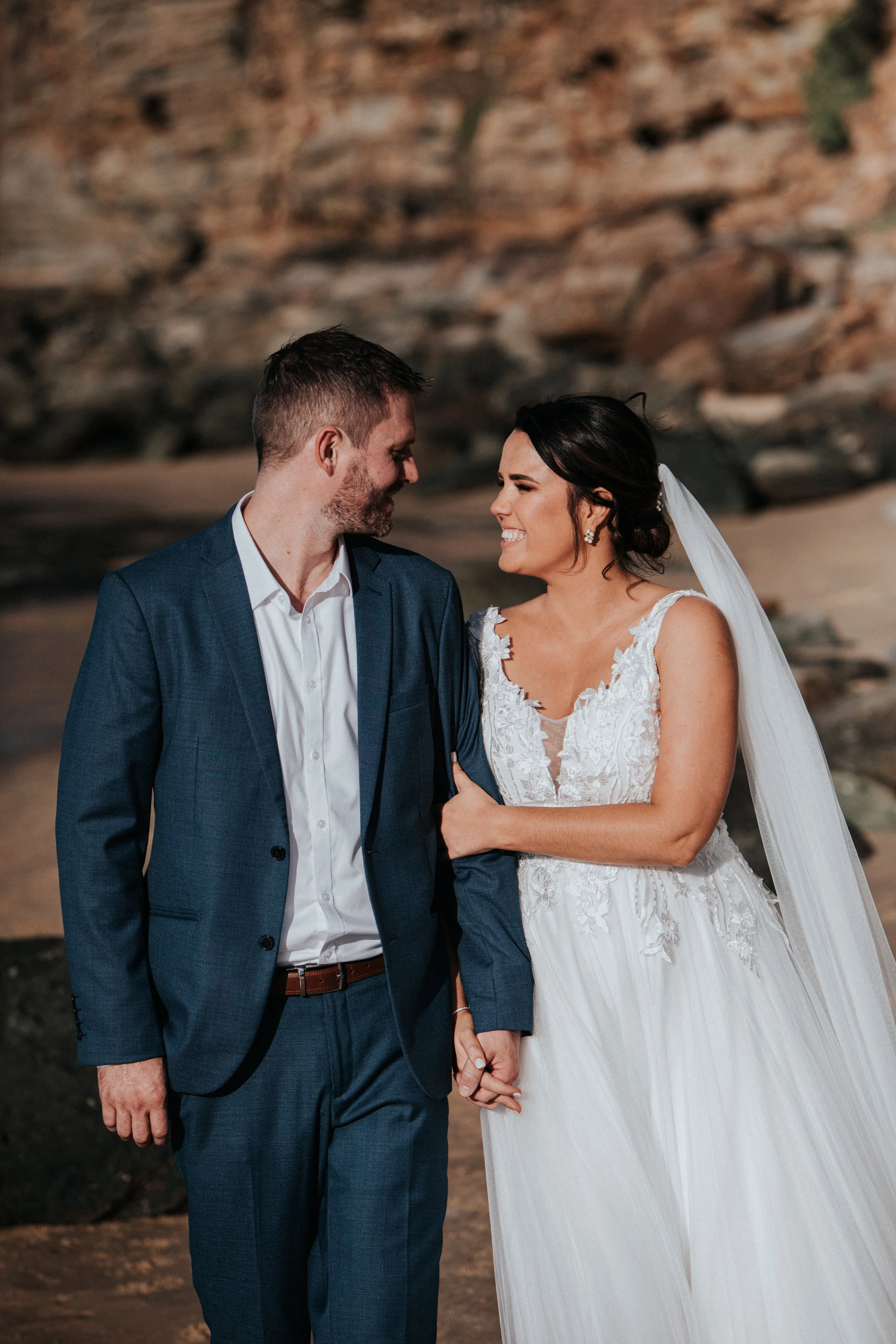 A bride and groom holding hands and smiling at each other on a beach with rocky cliffs in the background.