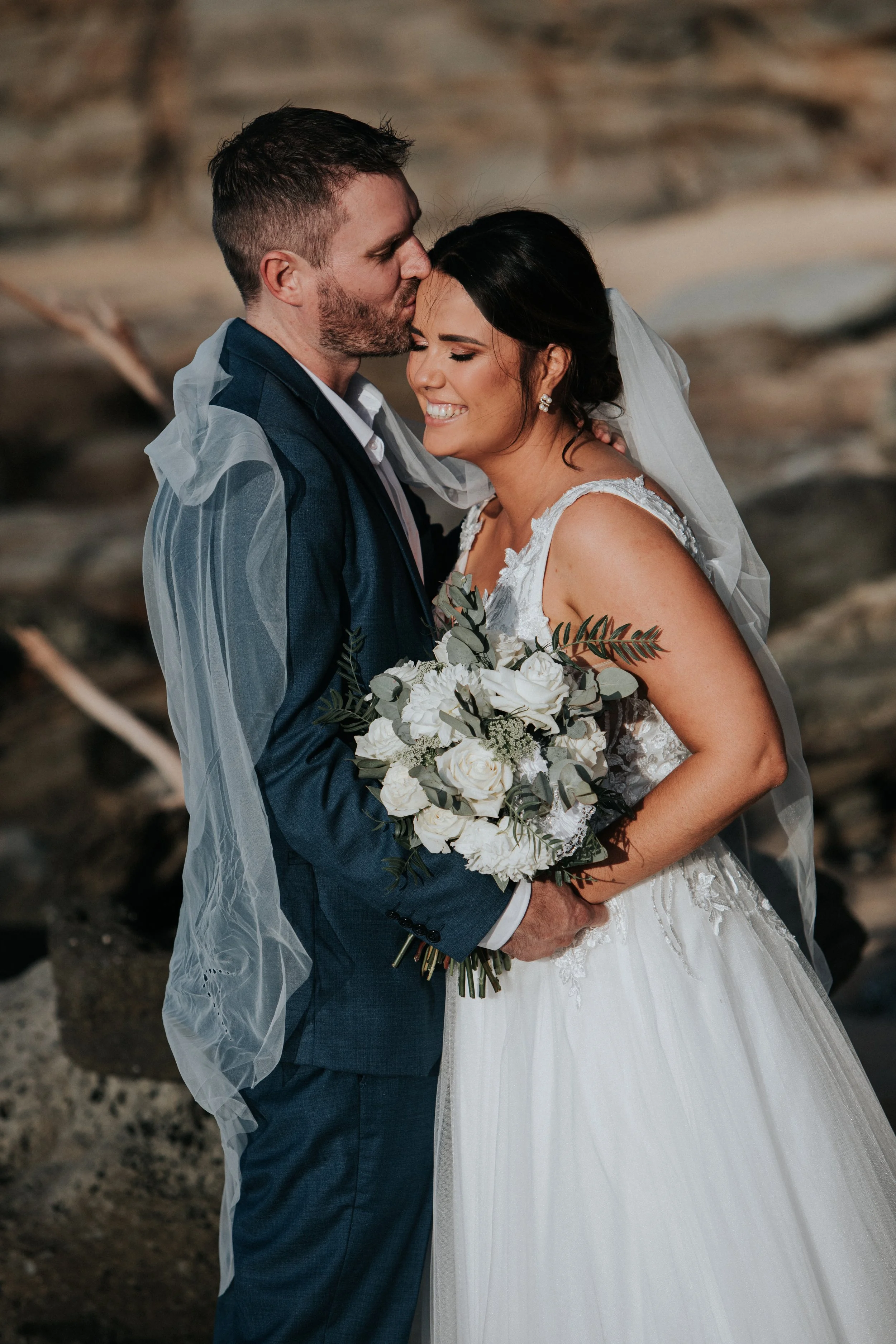 A newlywed couple sharing a tender moment on the beach, with the groom kissing the bride's forehead. The bride is holding a bouquet of white flowers and greenery, wearing a white lace wedding gown and veil. The groom is dressed in a navy suit, with a