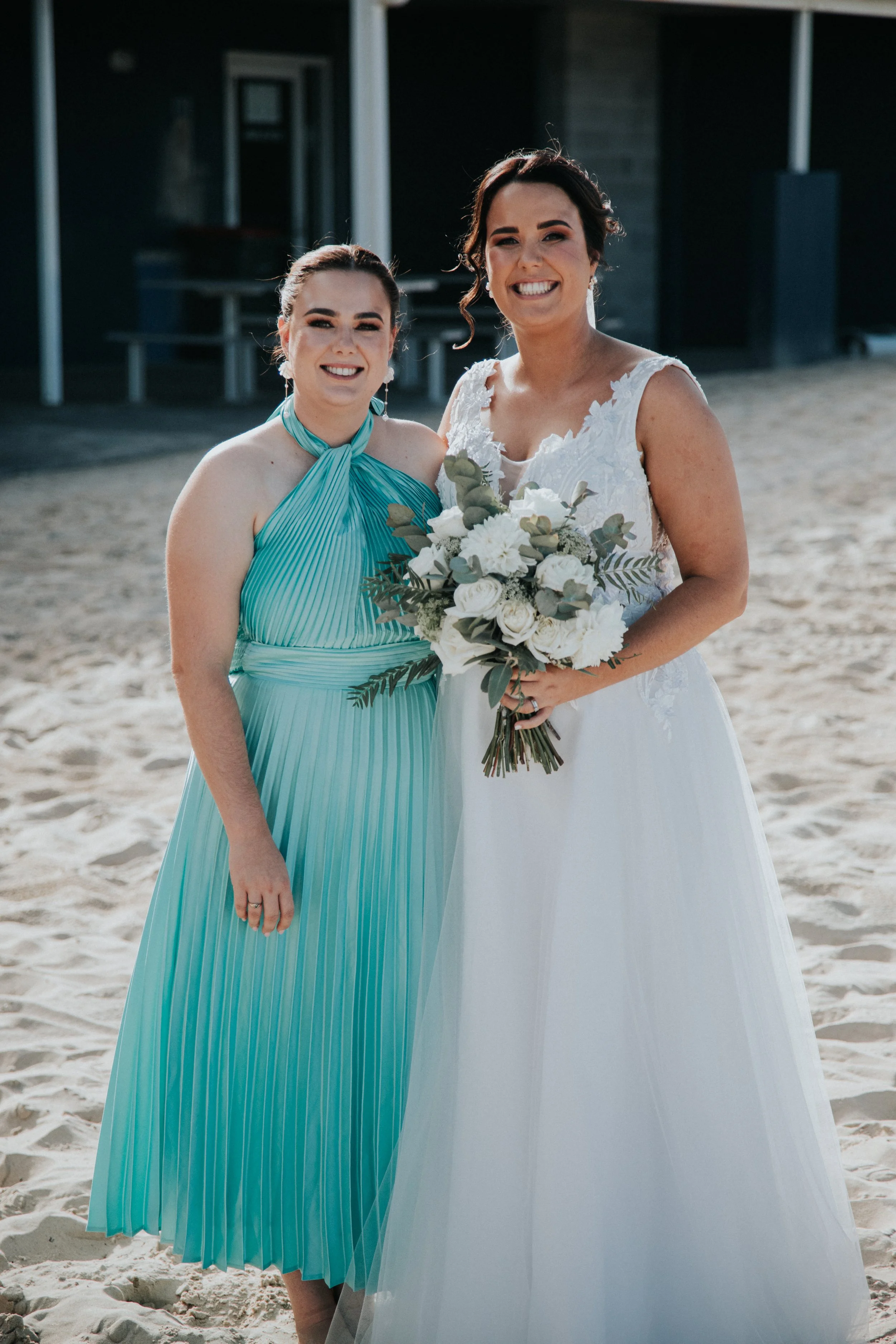 Two women in wedding attire standing on a sandy beach, smiling at the camera, with one holding a bouquet of white flowers and greenery.
