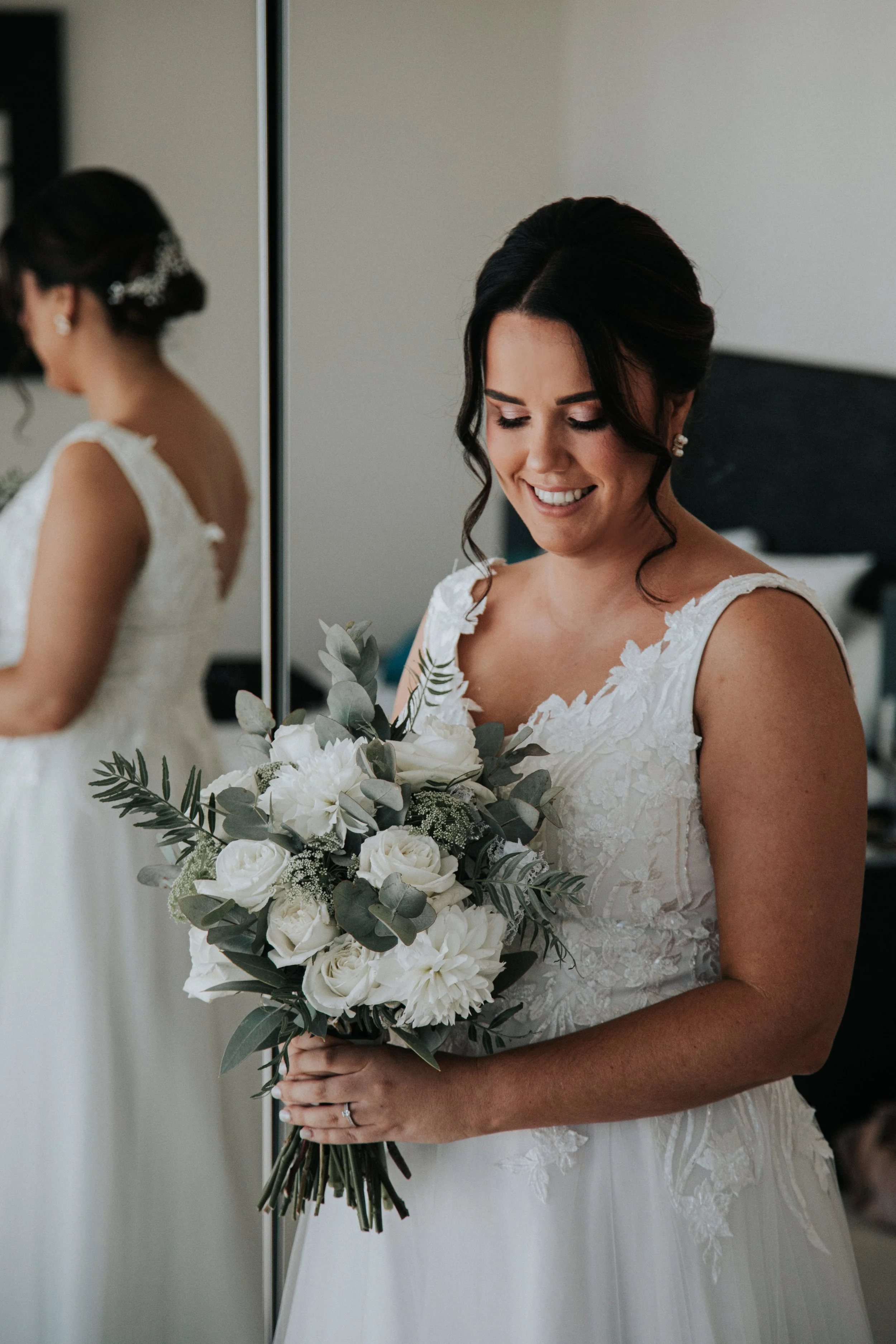 A bride in a white lace wedding dress holding a bouquet of white roses, dahlias, and greenery, smiling as she looks down.