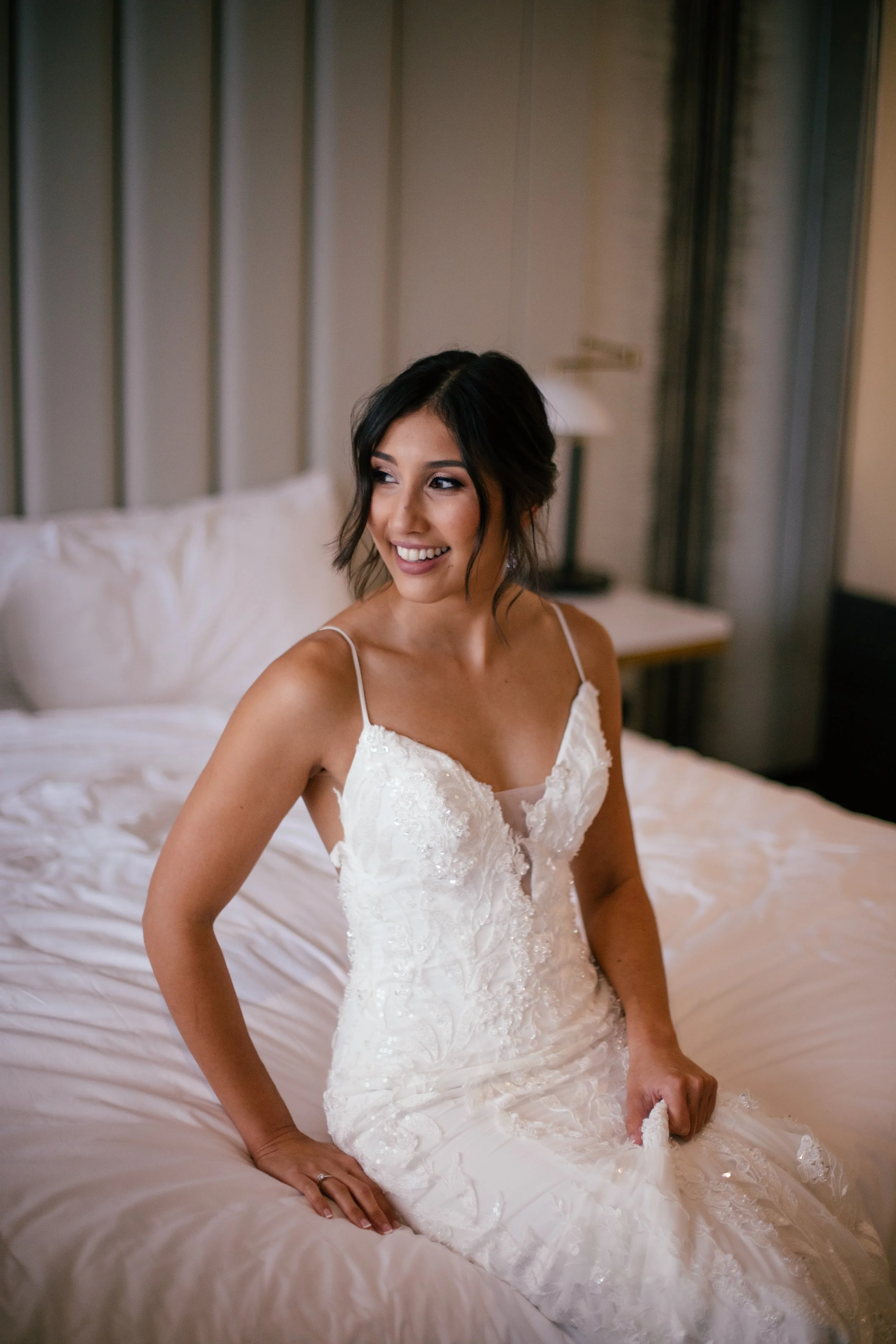 A smiling bride in a white wedding dress sitting on a bed in a hotel room.