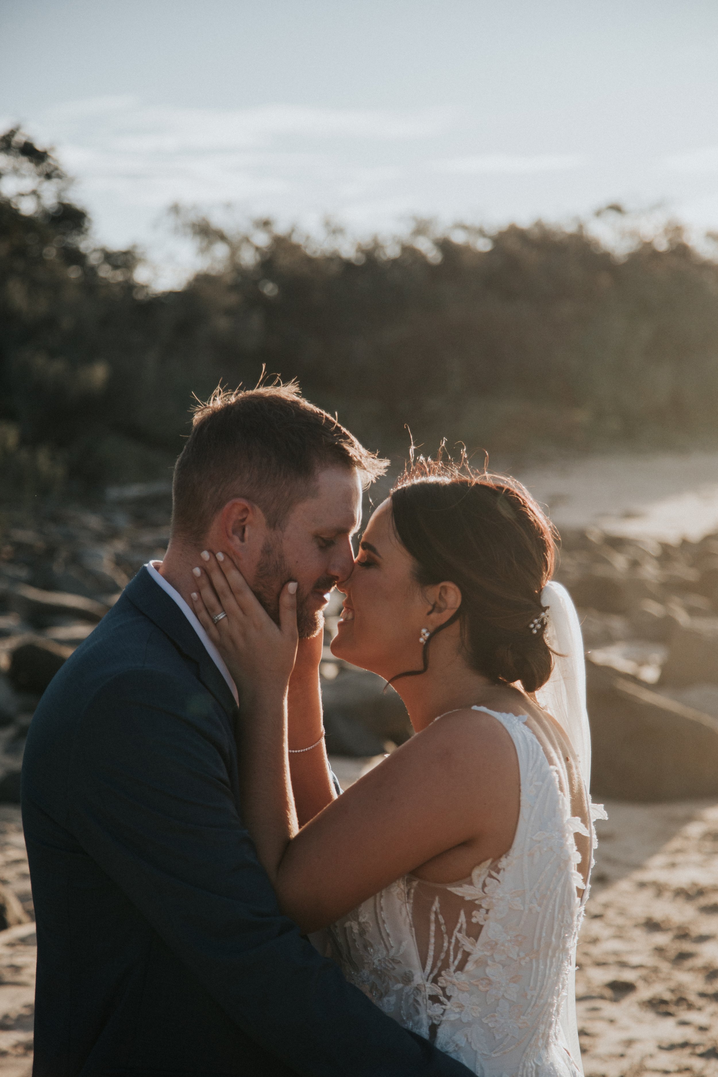 A bride and groom embrace on a beach at sunset, with the man in a dark suit and the woman in a white lace wedding dress, touching foreheads and holding each other's faces.