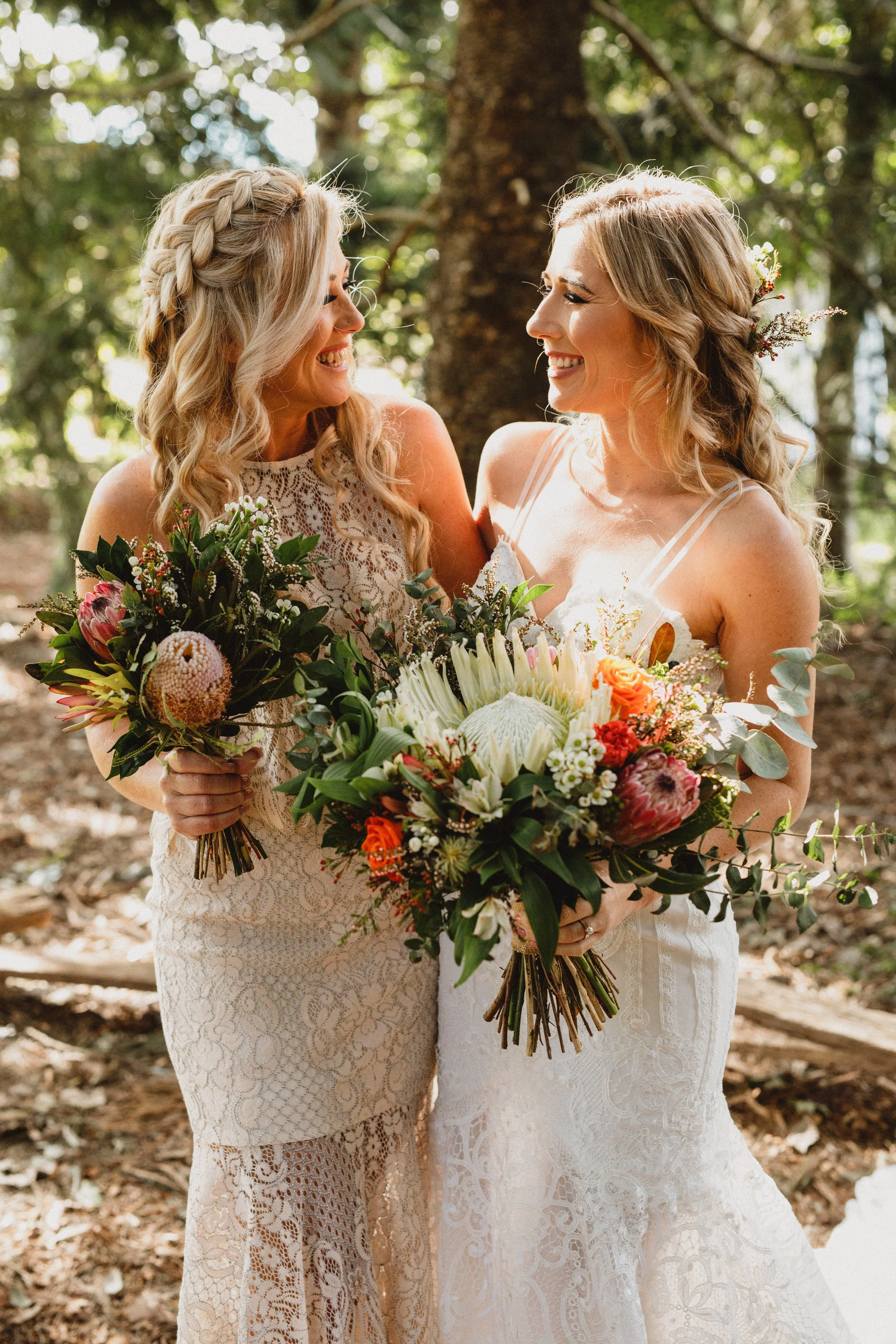 Two women in wedding dresses holding bouquets, smiling at each other in an outdoor wooded setting.