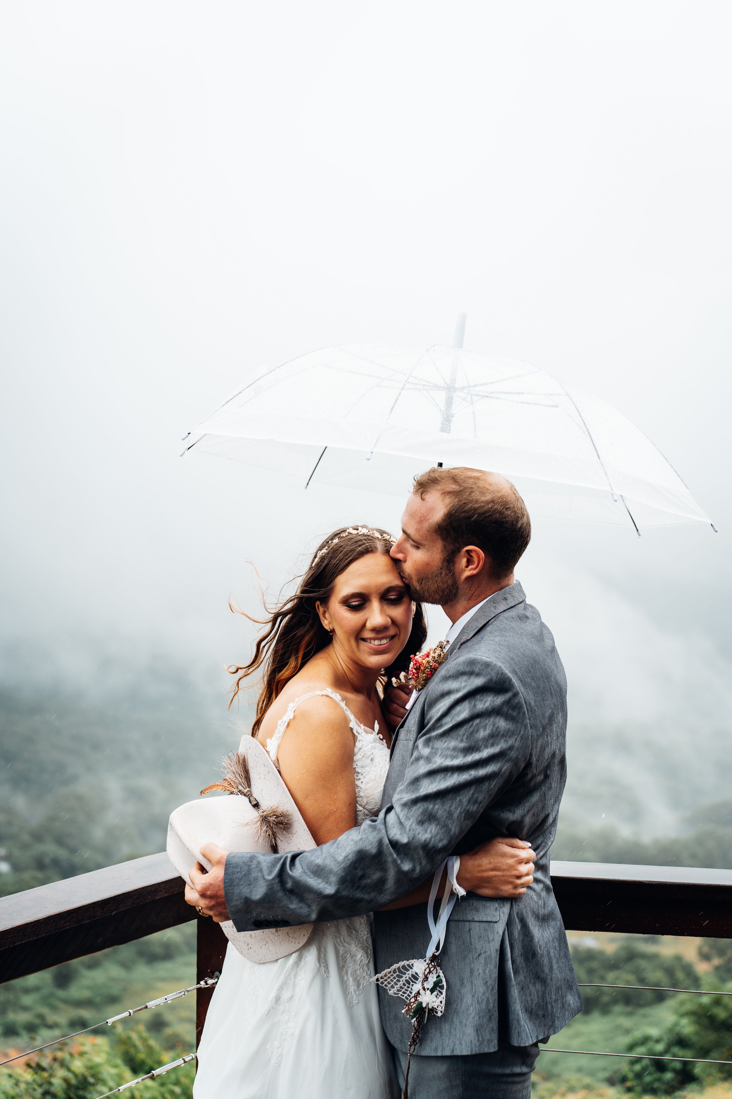 A newlywed couple stands close together under a clear umbrella on a foggy outdoor balcony, with trees and mist in the background, sharing an intimate moment.