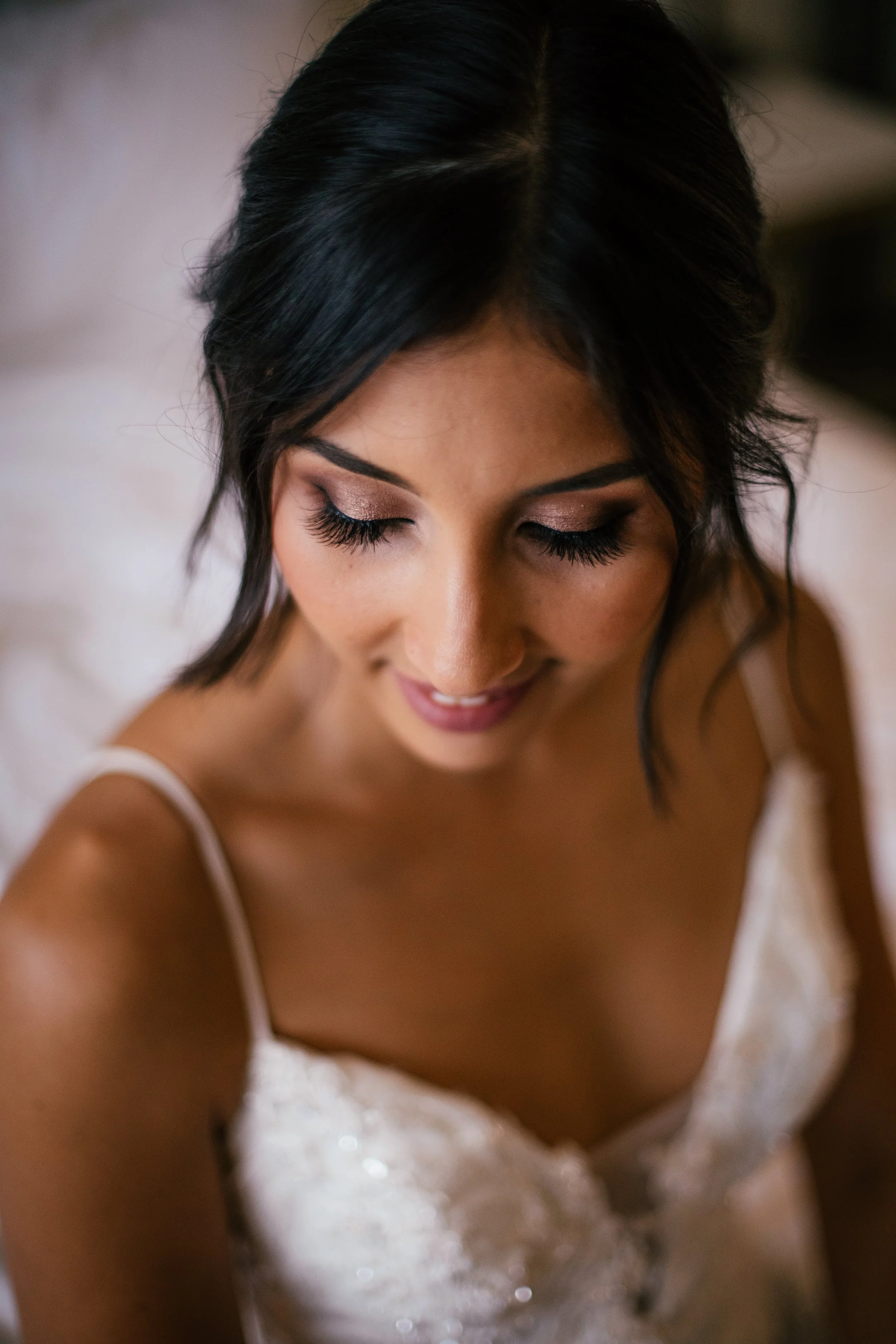 A woman with dark hair and long eyelashes wearing a white dress, smiling gently with her eyes closed.
