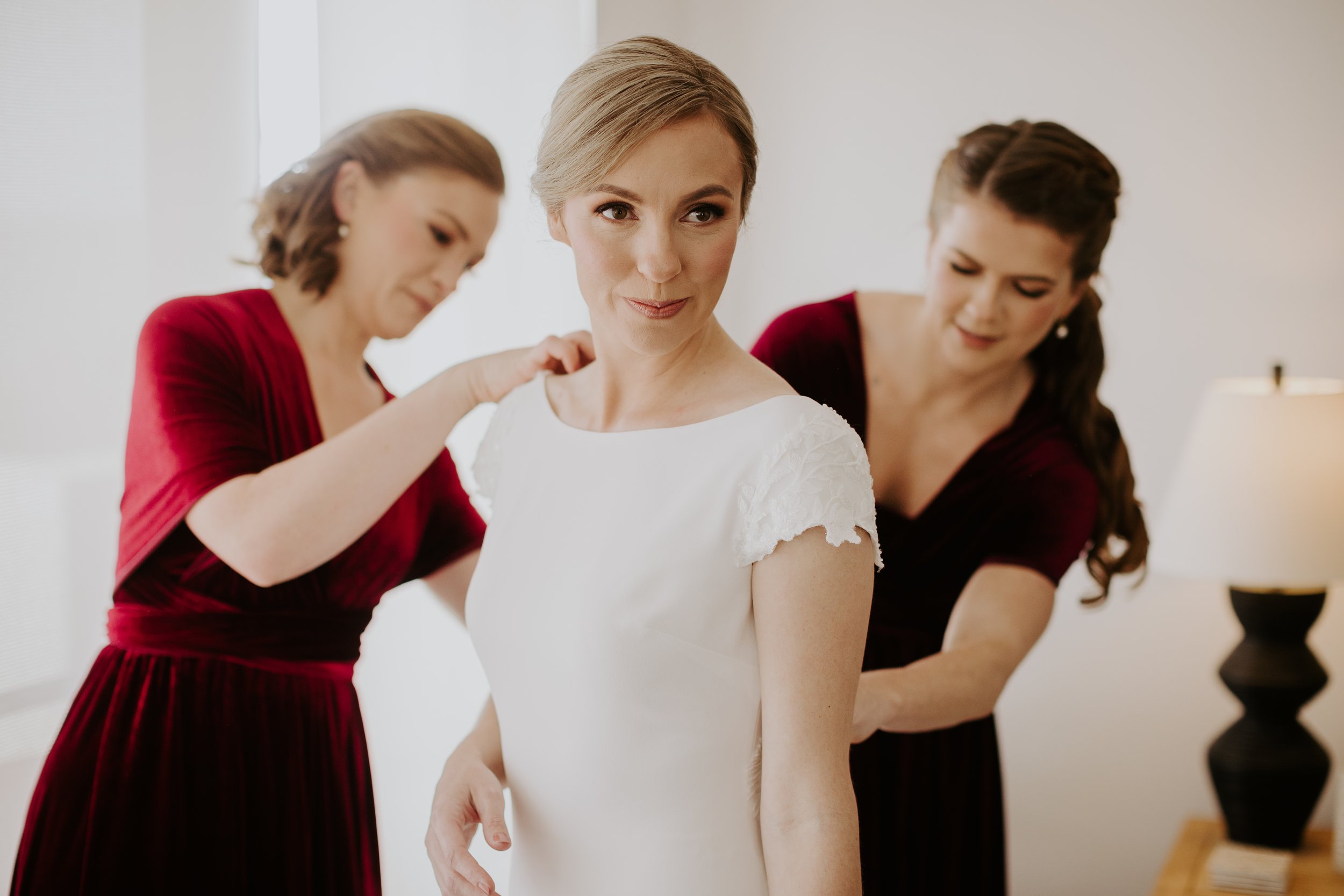 A bride in a white dress stands in front of two women helping her get ready, with one woman dressing her shoulders and the other adjusting her dress. The room has neutral walls and a lamp in the background.