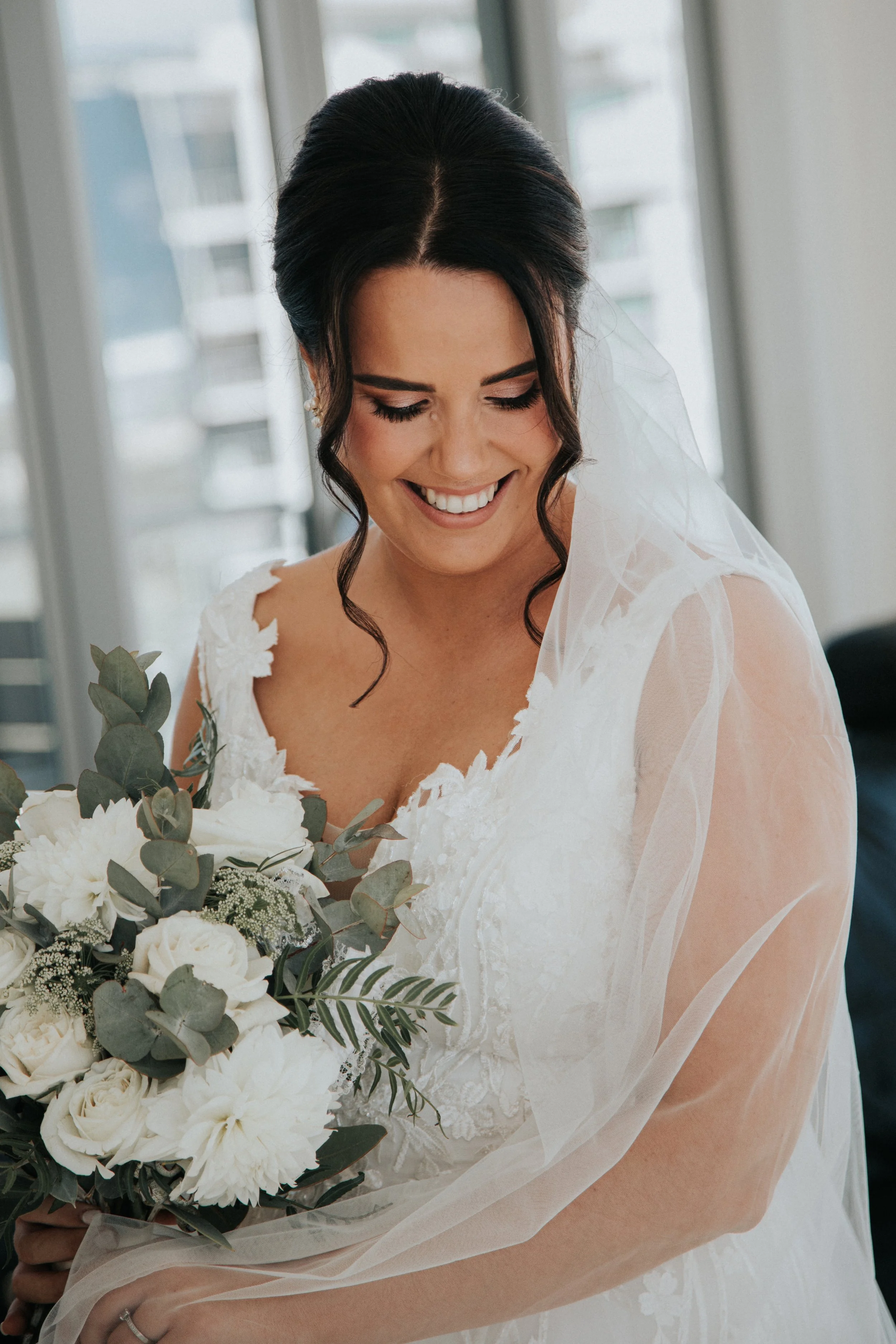 A bride in a white wedding dress with lace details, smiling and holding a bouquet of white flowers and greenery, with soft natural light coming through a window behind her.