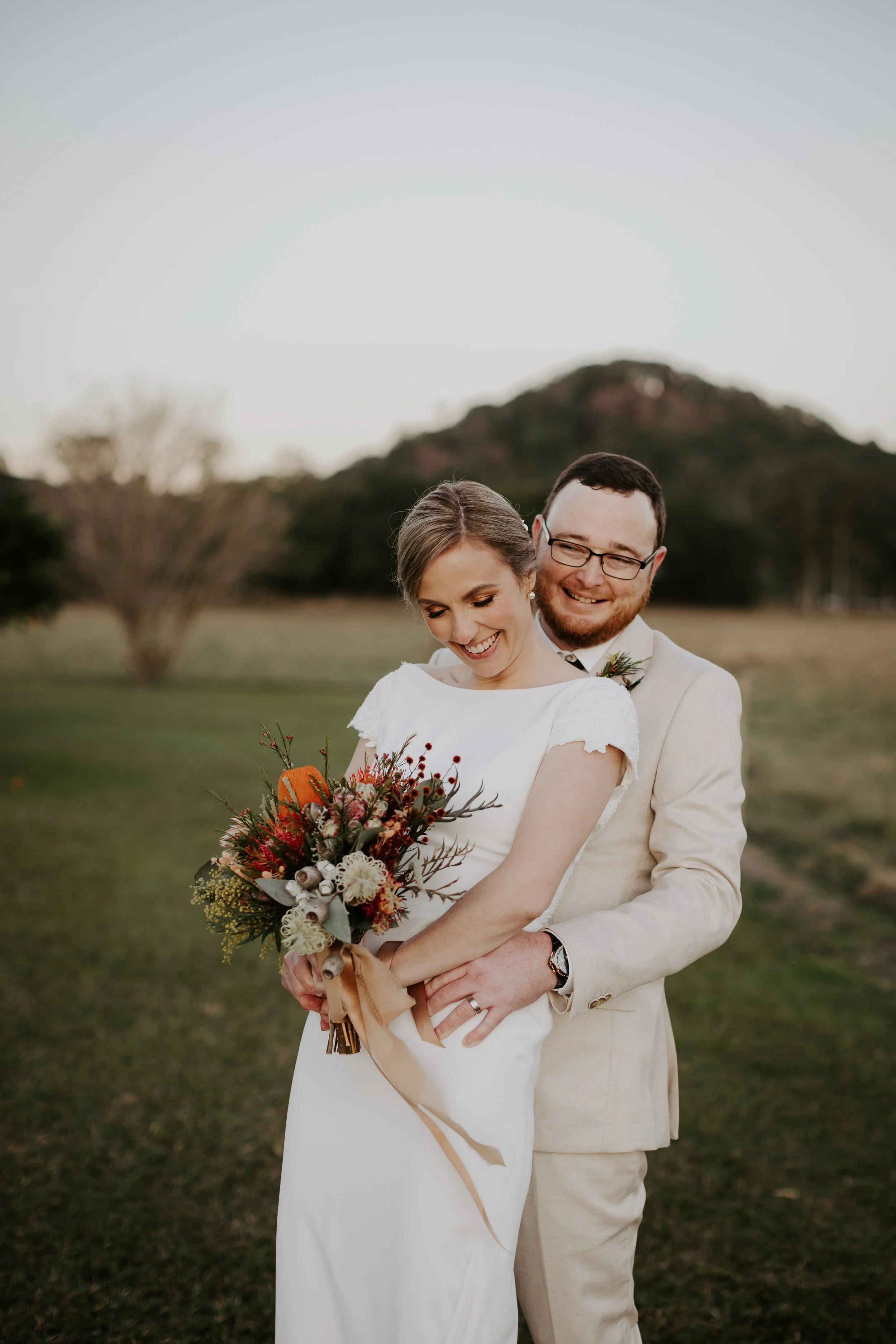 Bride and groom smiling and embracing outdoors at sunset, with the bride holding a bouquet of flowers.