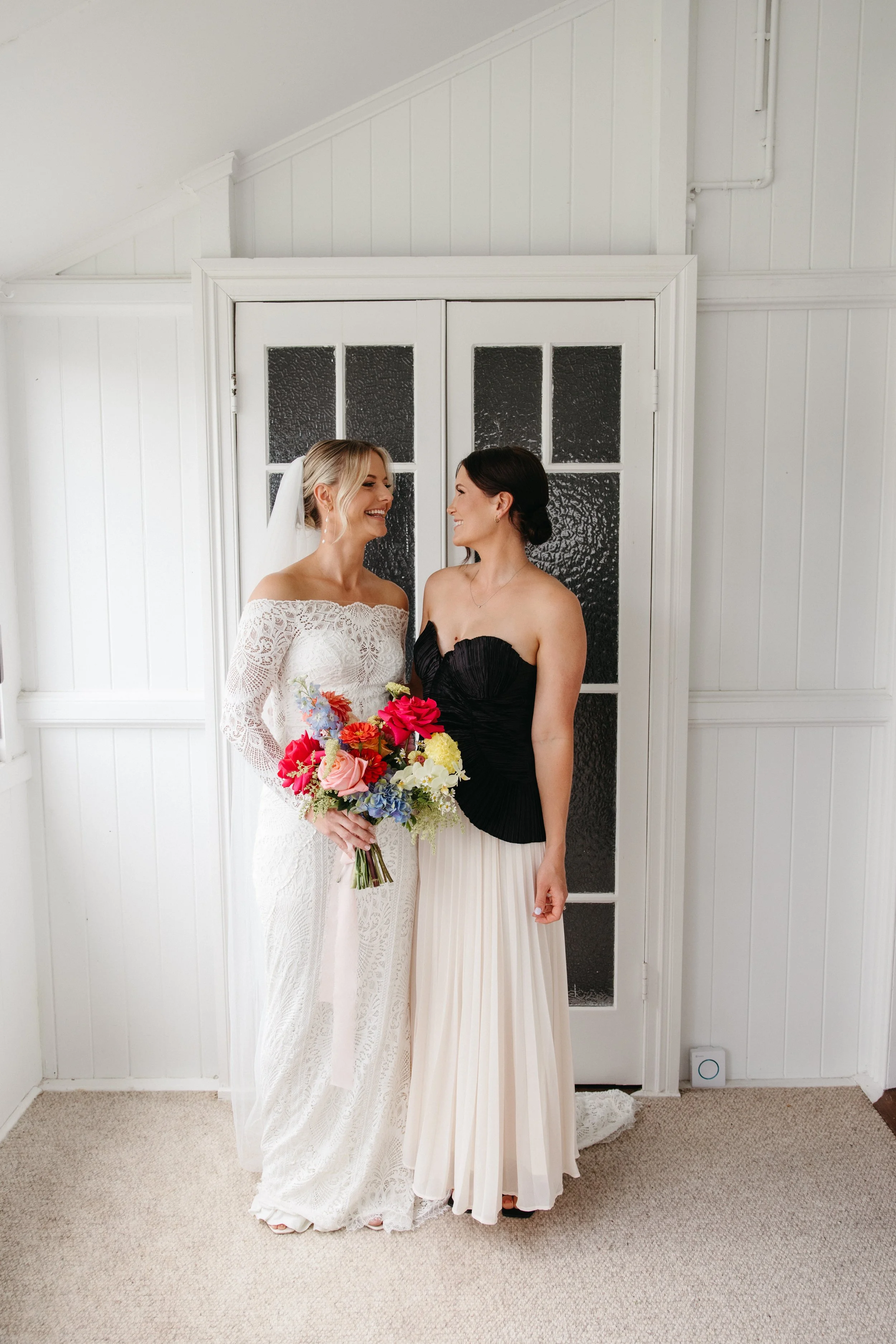 Two women, one in a white lace wedding dress holding a colorful bouquet, the other in a black strapless top and cream pleated skirt, smiling at each other in front of a white paneled door with textured glass panels.