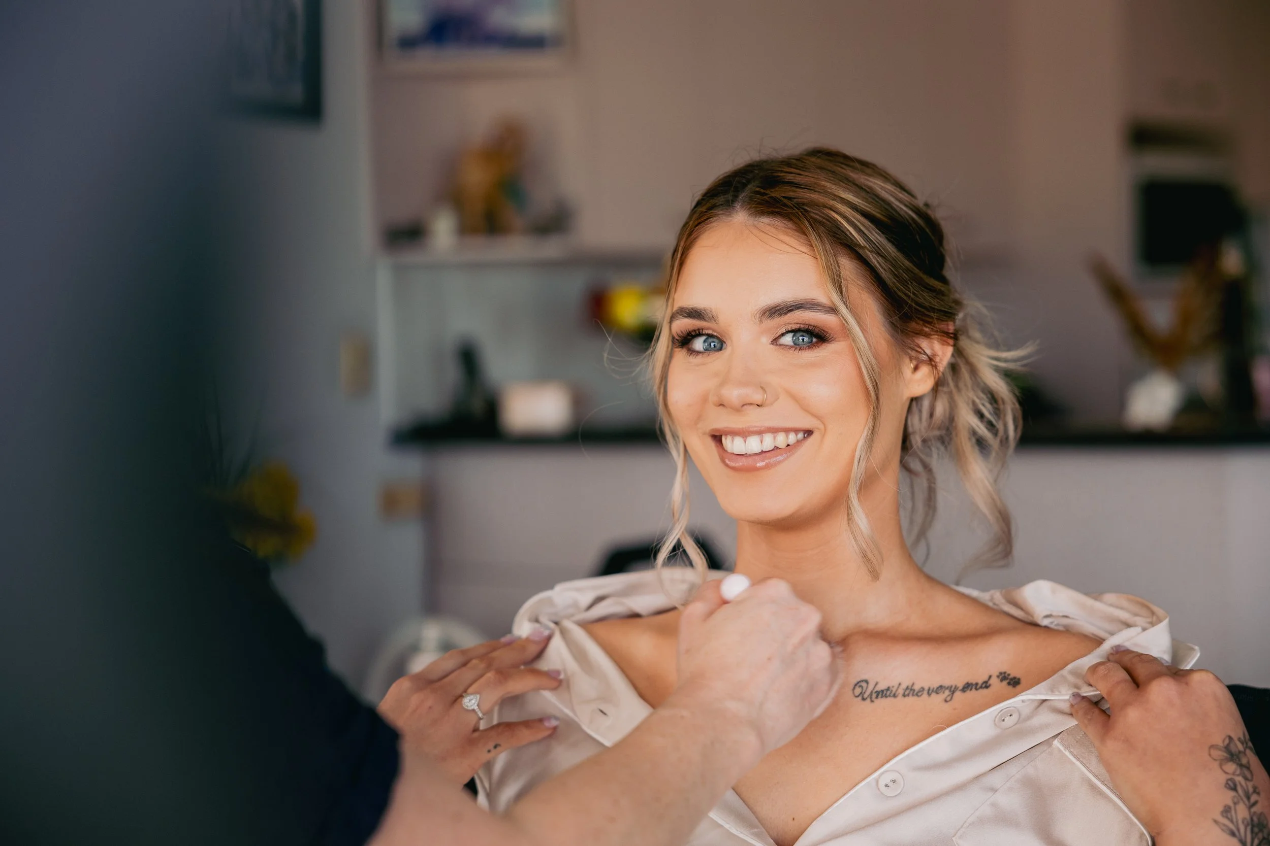 A smiling woman with blonde hair and blue eyes getting her shirt adjusted by another person in a cozy indoor setting.