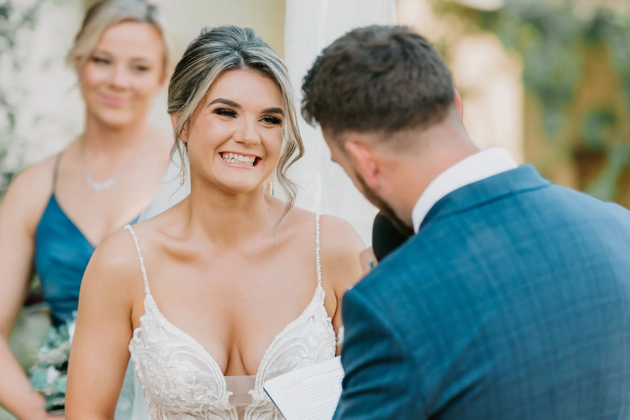 Bride smiling at groom during wedding ceremony, officiant holding a book in the foreground, bridesmaid in the background, outdoor setting with trees.