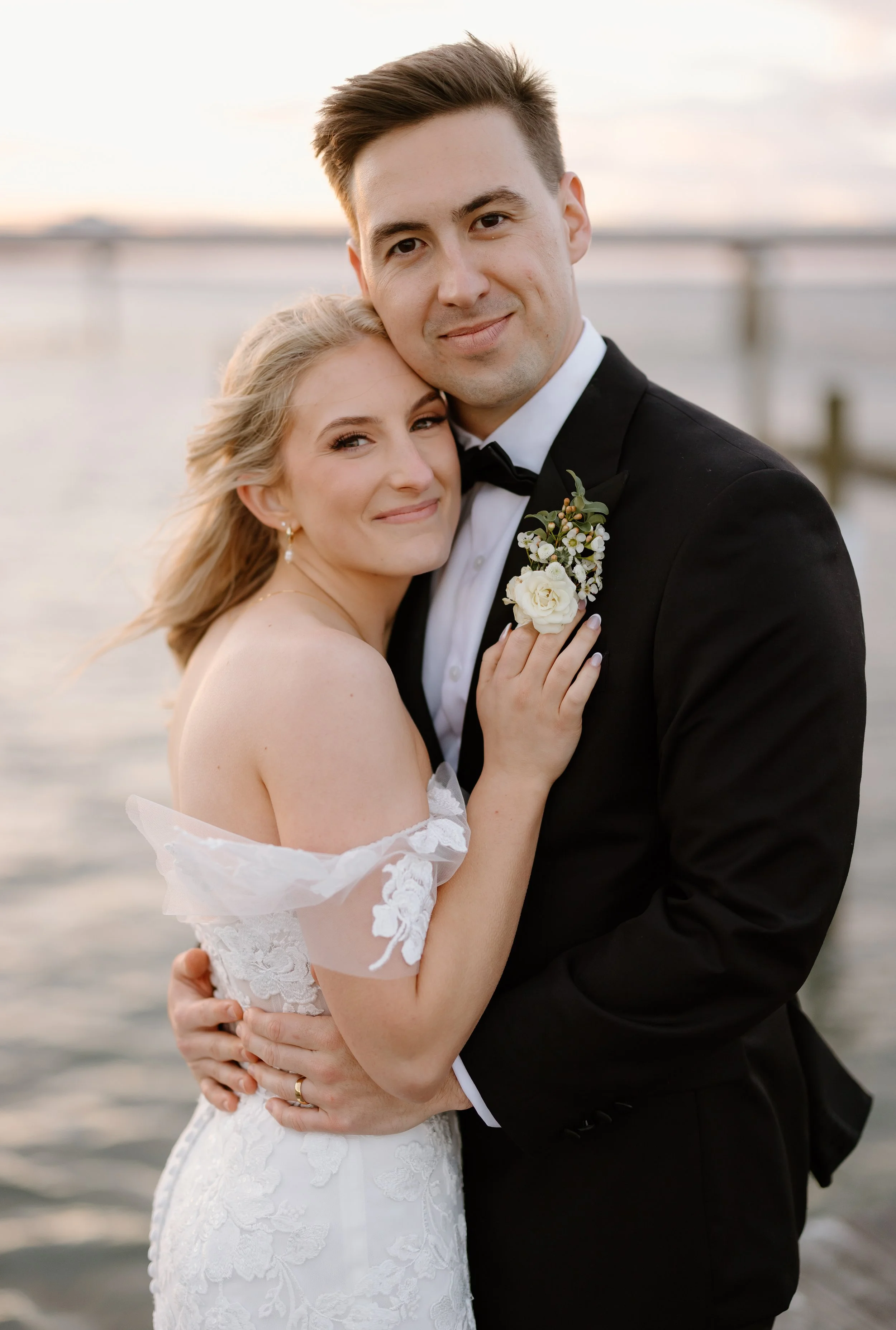 A newlywed couple embracing by the water at sunset, with the bride in a lace wedding dress and the groom in a black tuxedo.