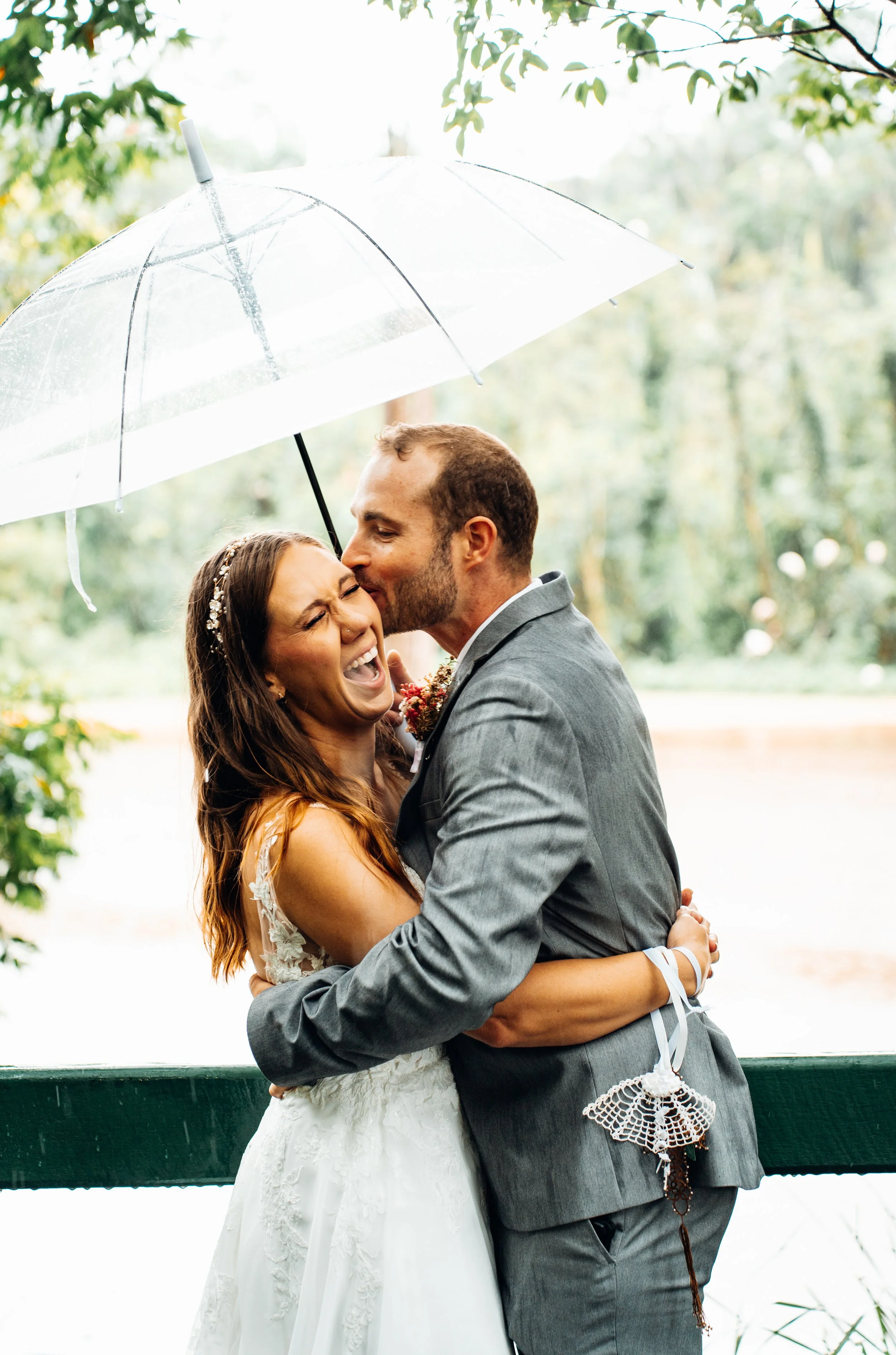 A bride and groom embracing under a clear umbrella, with the groom kissing the bride on the forehead, outdoors near a green railing and trees.