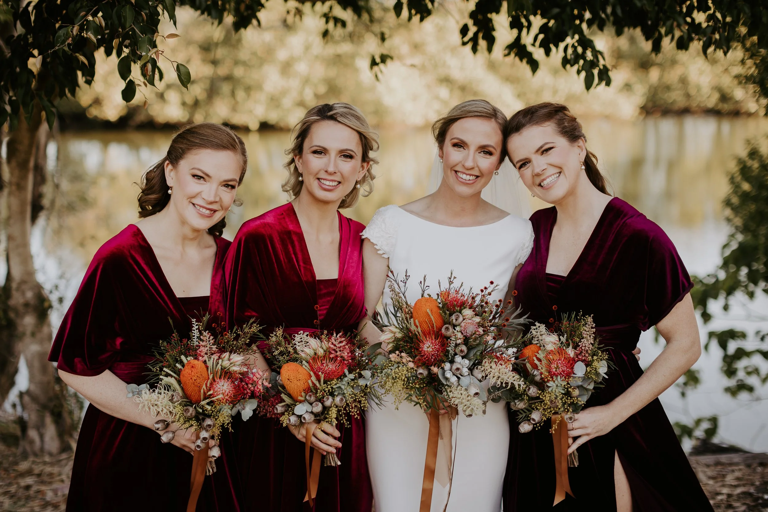 Four women, one in a white wedding dress and three in deep red velvet dresses, standing outdoors near a body of water, holding colorful bouquets, smiling, with trees and autumn foliage in the background.