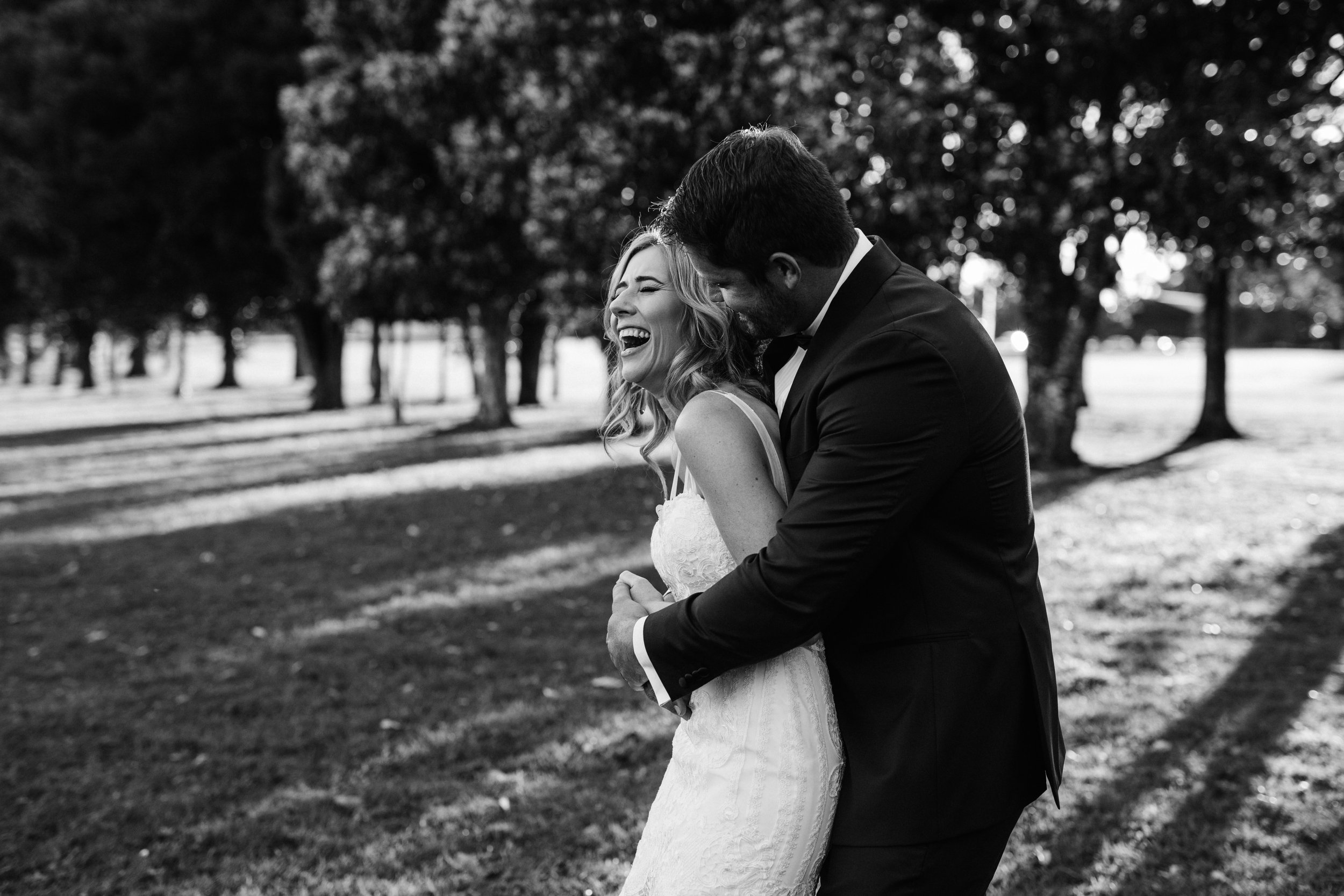 A black and white photo of a bride and groom laughing and dancing outdoors in a park, with trees in the background.