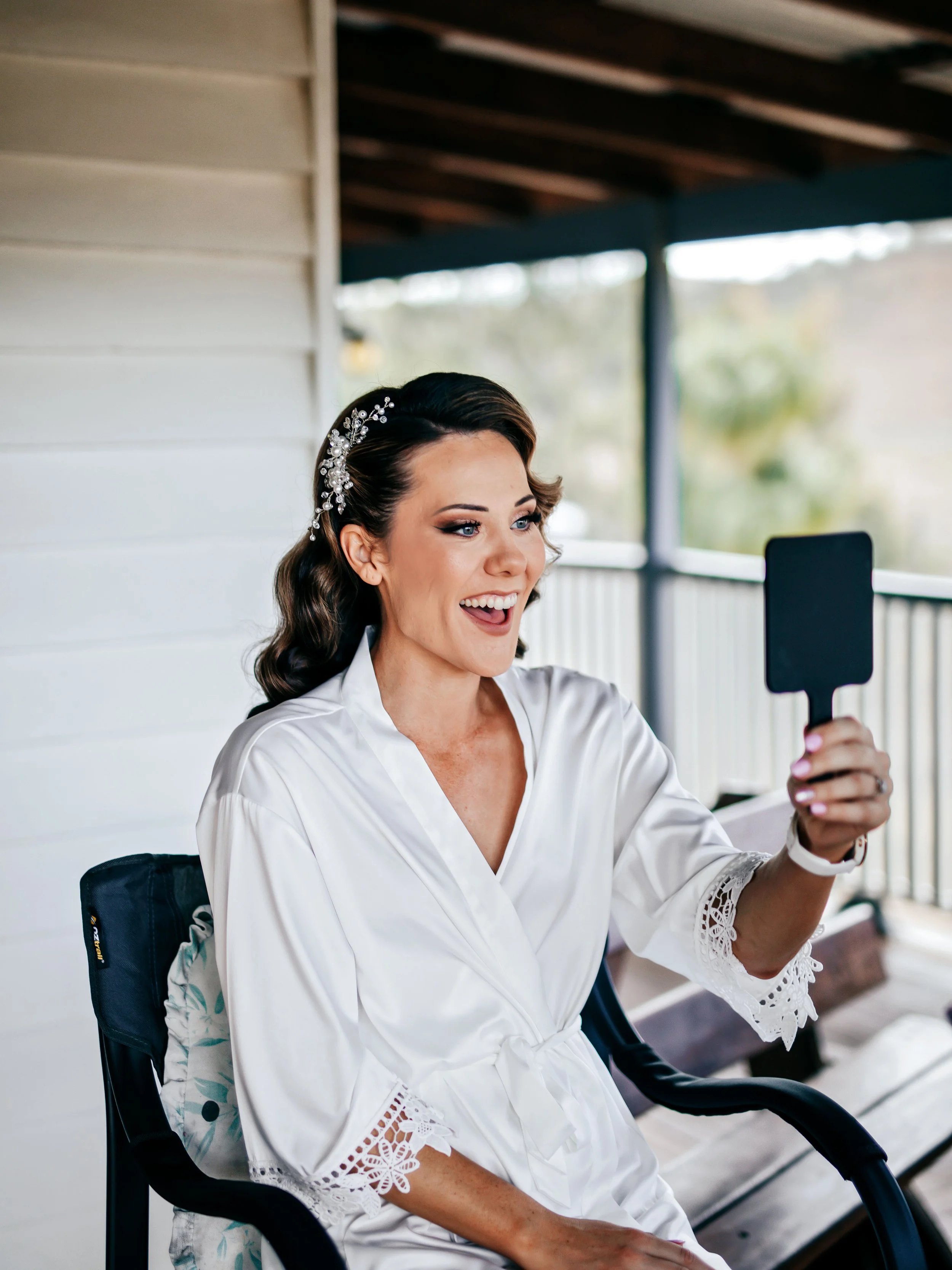 A woman with dark hair styled in loose waves, wearing a white robe with lace details, is sitting on a chair on a porch. She is smiling and holding a black handheld mirror, looking at her reflection.