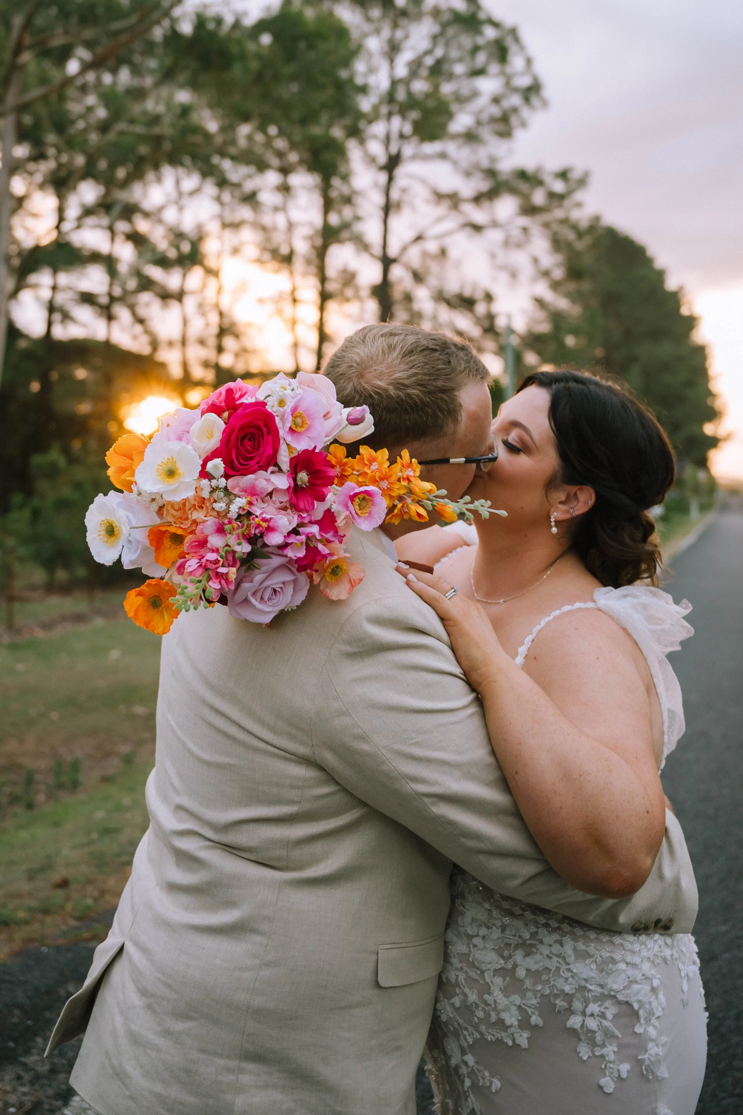 A newlywed couple sharing a kiss outdoors during sunset. The bride is holding a bouquet of vibrant pink, orange, and white flowers. The background features trees and a setting sun.