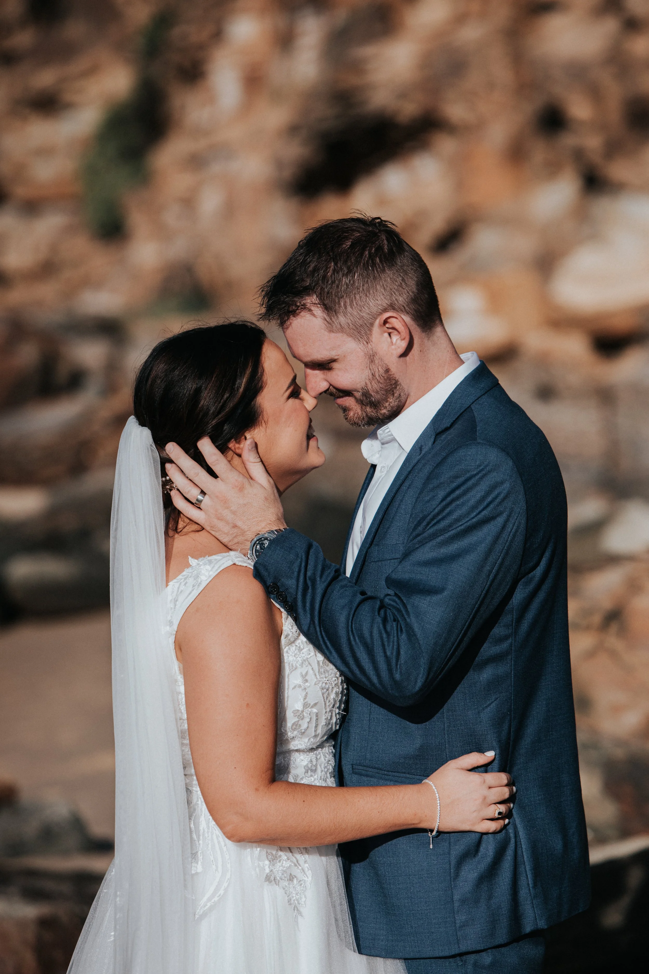 A bride and groom sharing an intimate moment outdoors, holding each other close with their foreheads touching, against a rocky background.