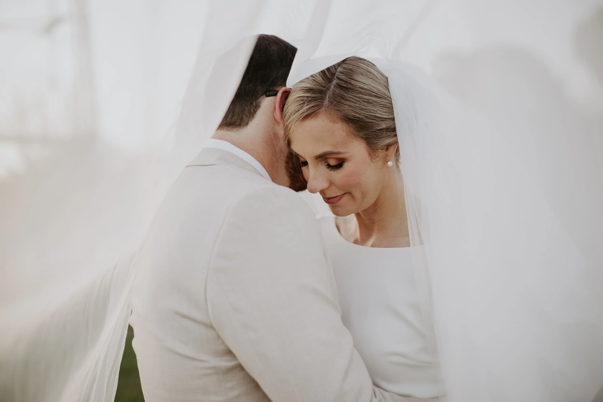 A bride and groom standing closely together, smiling gently, under a white veil during their wedding.