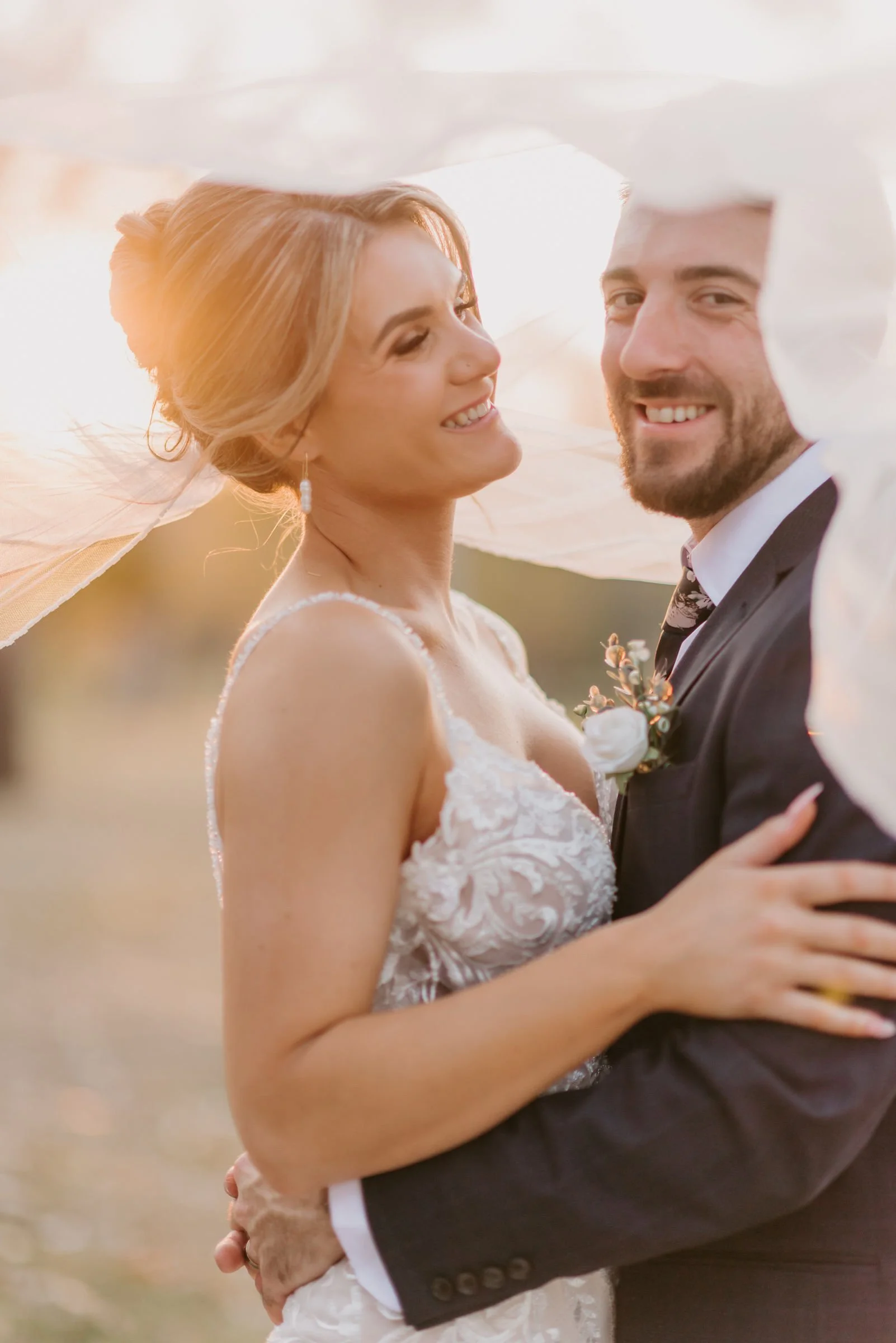 A newlywed couple smiling and hugging outdoors, with a sunset background.