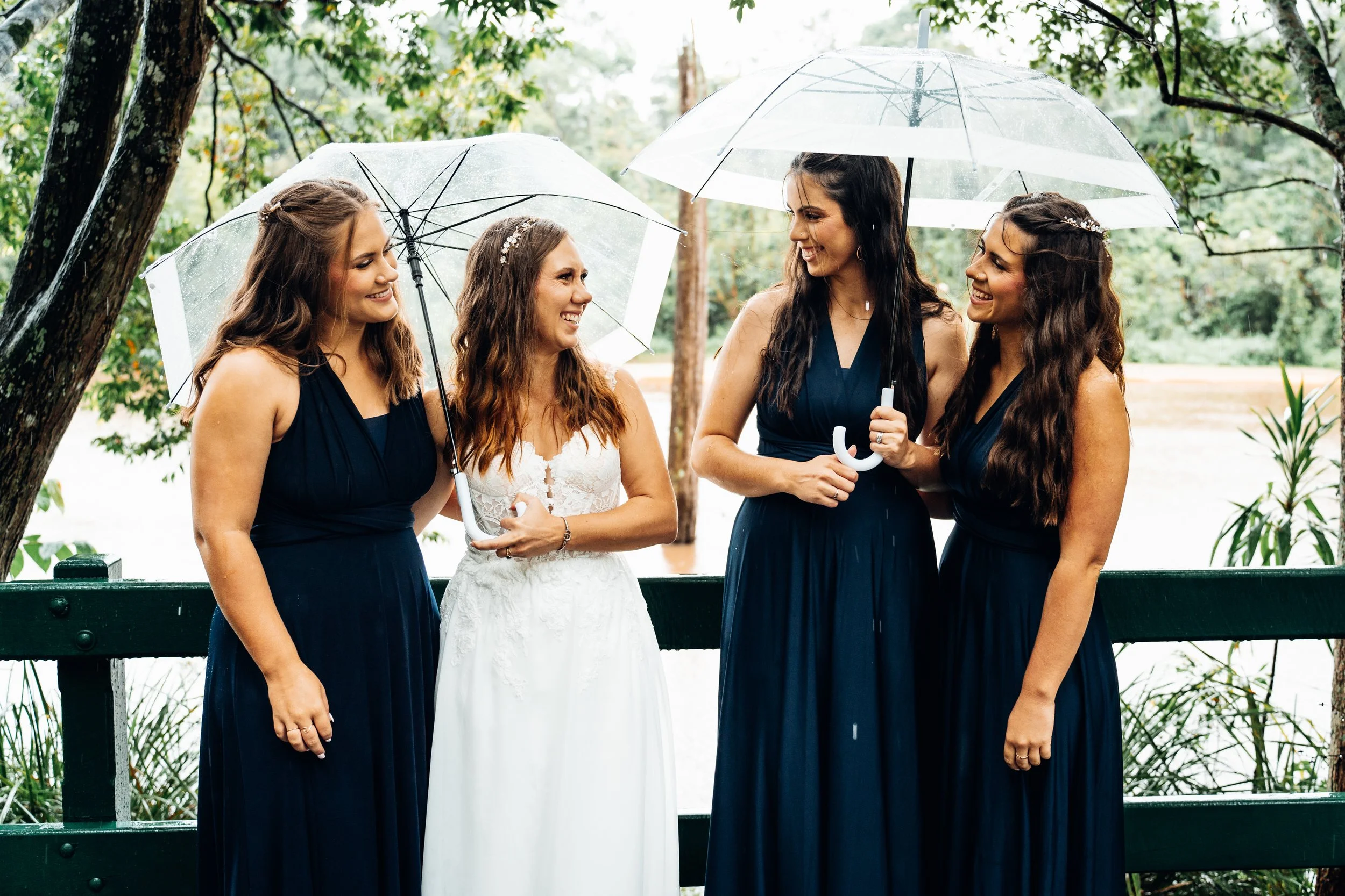 Four women, including a bride in a white gown, sharing a moment outside with umbrellas during a rainy day.