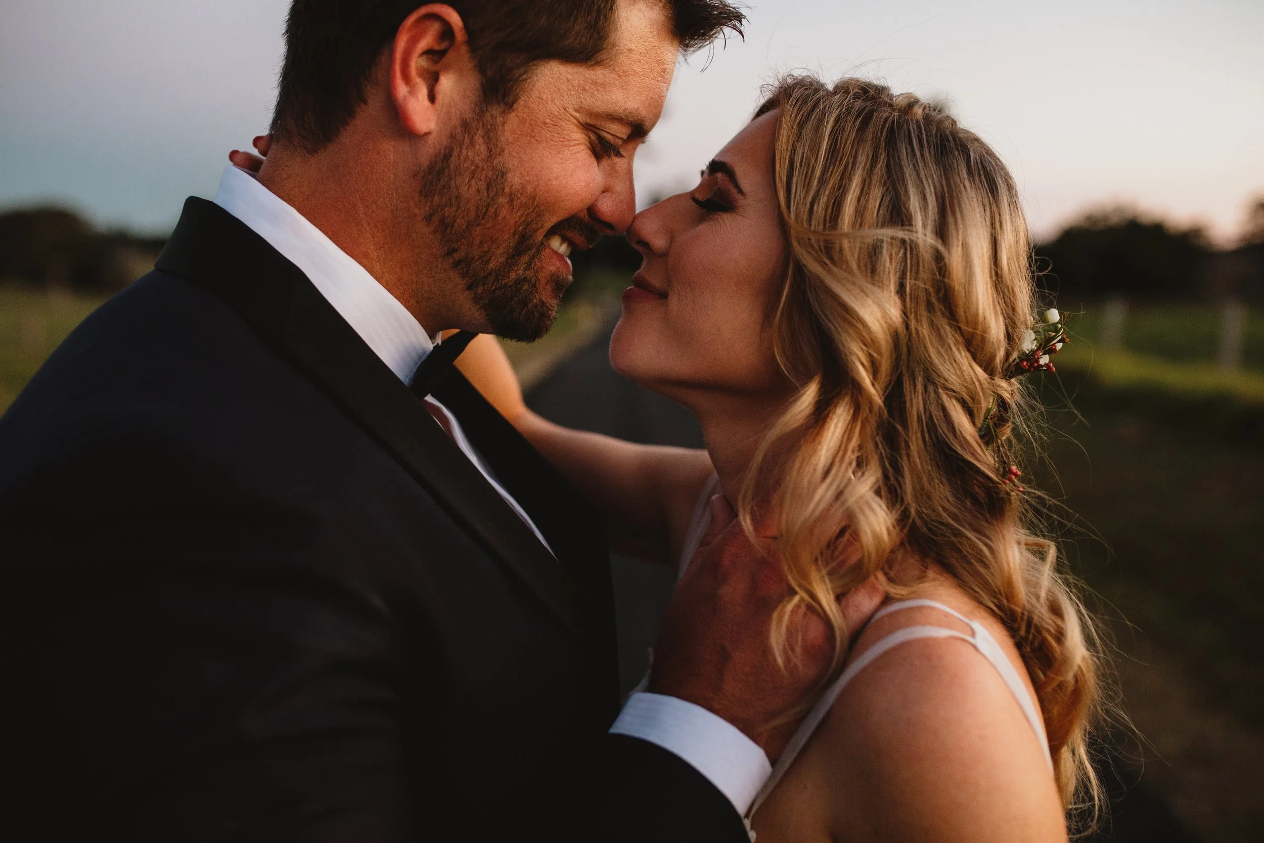 A couple is close together, nose to nose, smiling with their eyes closed outdoors at sunset or dusk.