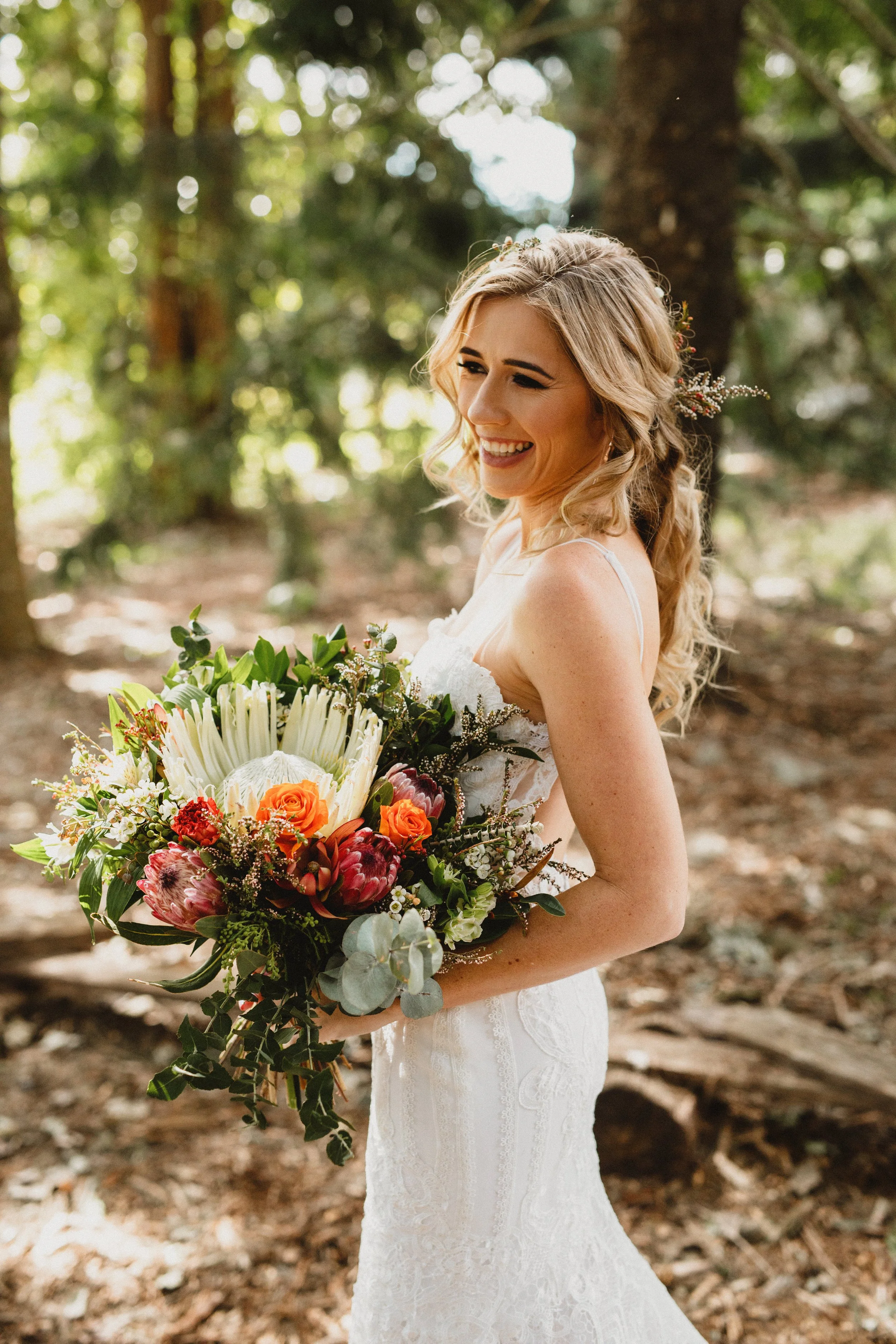 A woman in a white wedding dress holding a bouquet of flowers, smiling in a wooded outdoor setting.