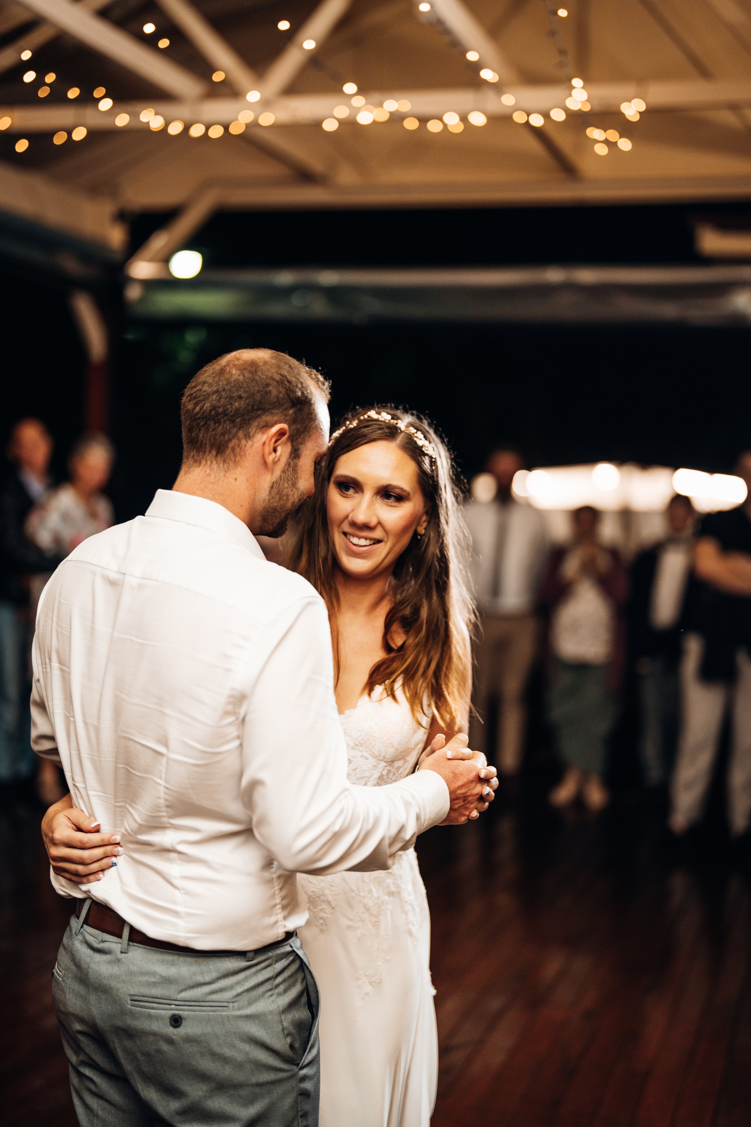 A bride and groom dancing at their wedding reception in a decorated venue with string lights, surrounded by guests.