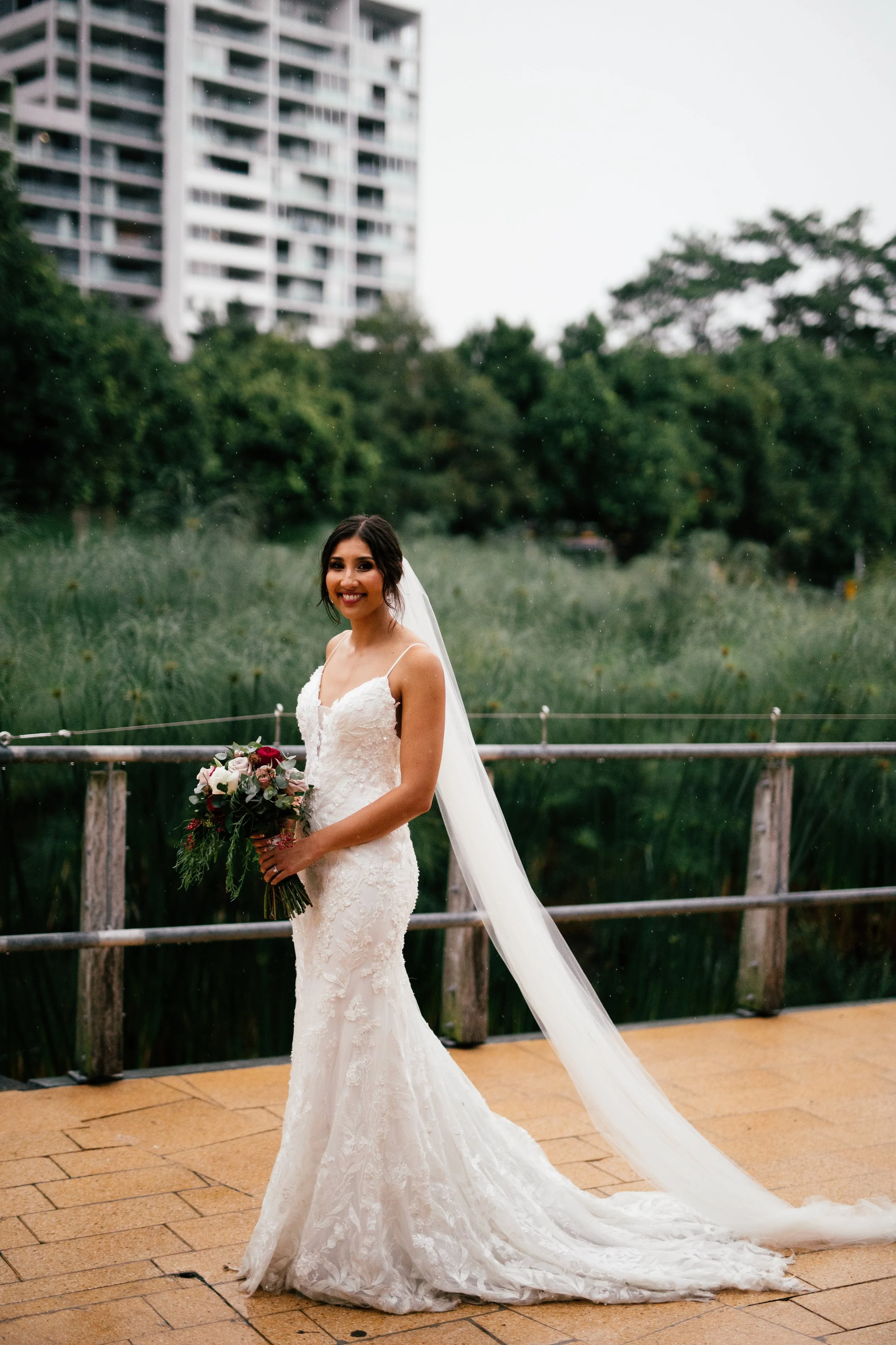A smiling bride standing outdoors on a wooden walkway, holding a bouquet of flowers, wearing a white wedding gown with a long veil, in front of lush greenery and a tall building in the background.