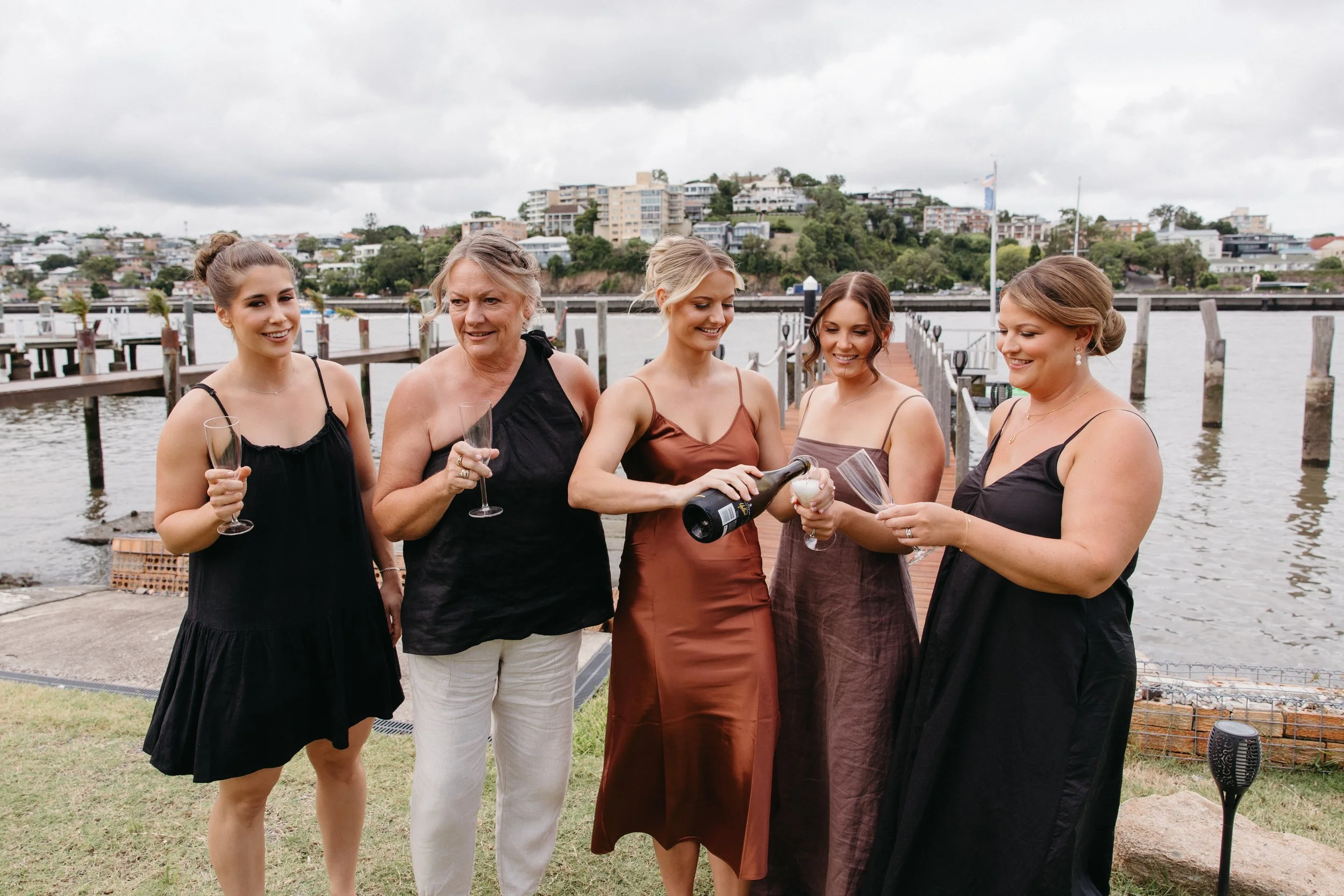 Five women celebrating outdoors by a waterfront, with one pouring champagne into a glass of another woman, all smiling and dressed in elegant dresses.