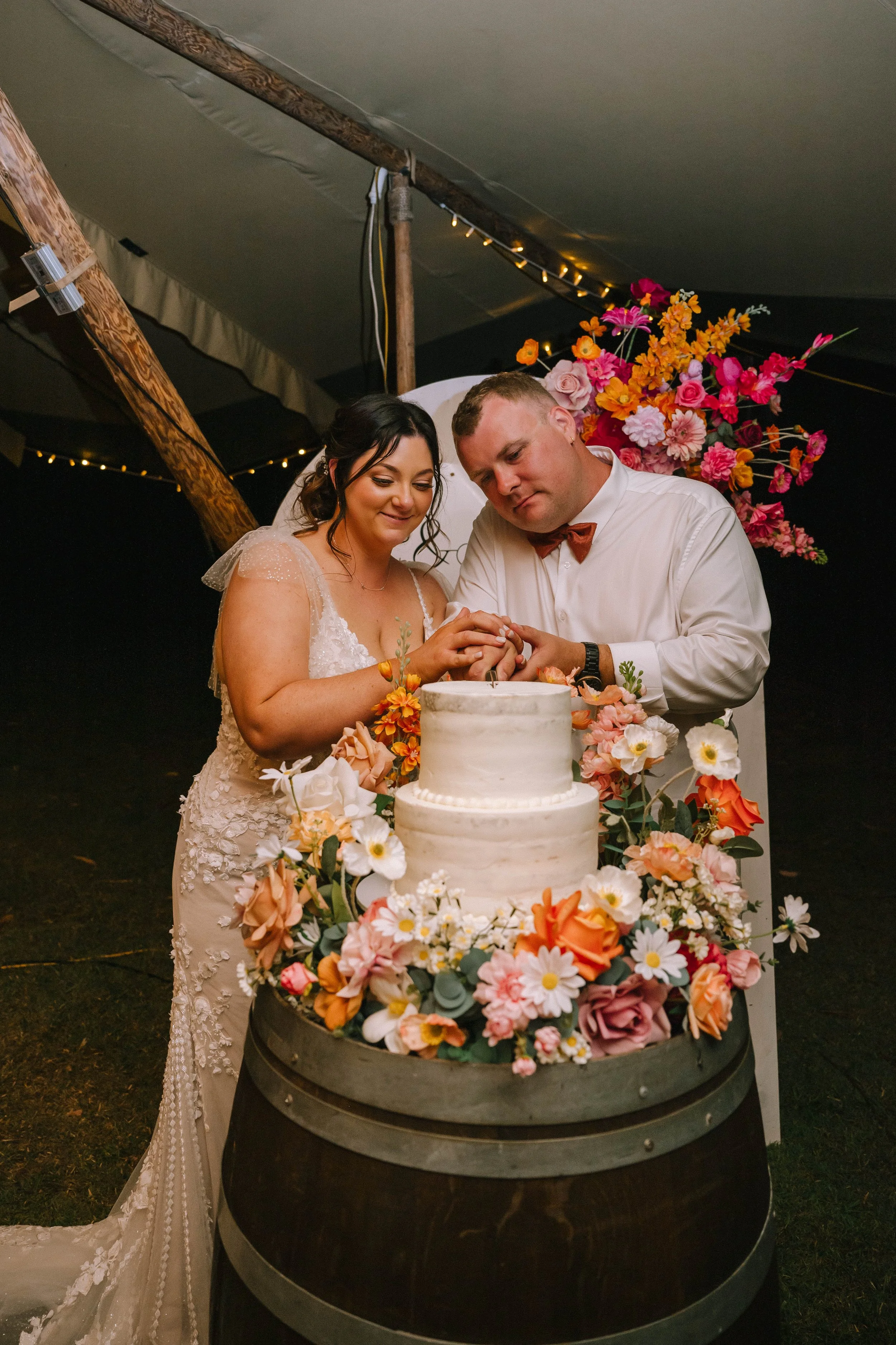 Bride and groom cutting wedding cake decorated with flowers inside a tent.