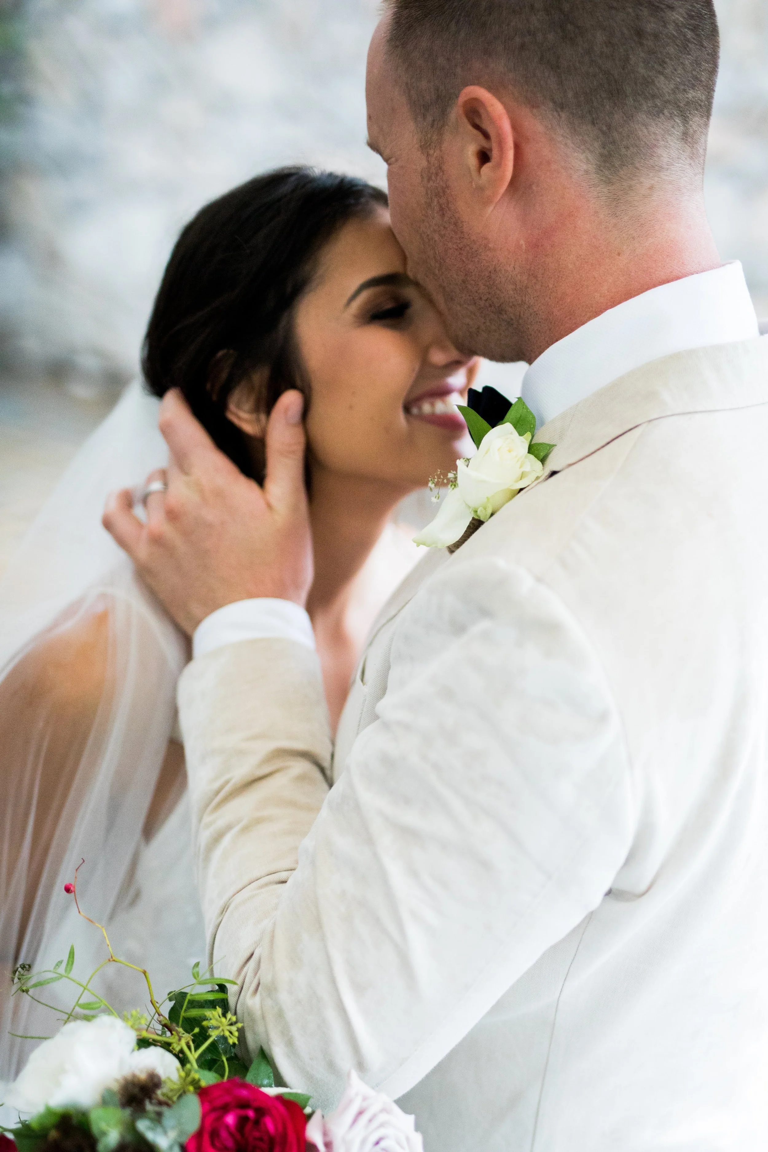 Close-up of a bride and groom with their faces touching, smiling, during their wedding, with the groom holding the bride's face and a bouquet of flowers visible in the foreground.