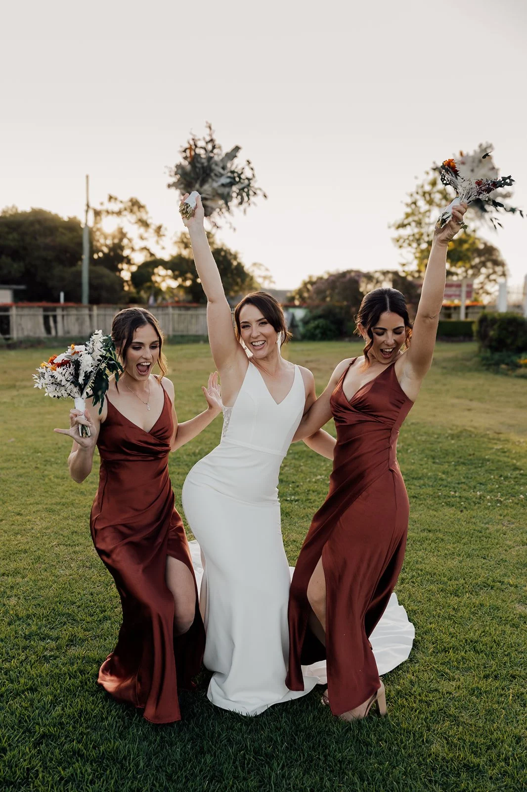 A bride in a white wedding gown celebrating with two bridesmaids in rust-colored dresses, outdoors on a grassy field during sunset, holding bouquets and smiling