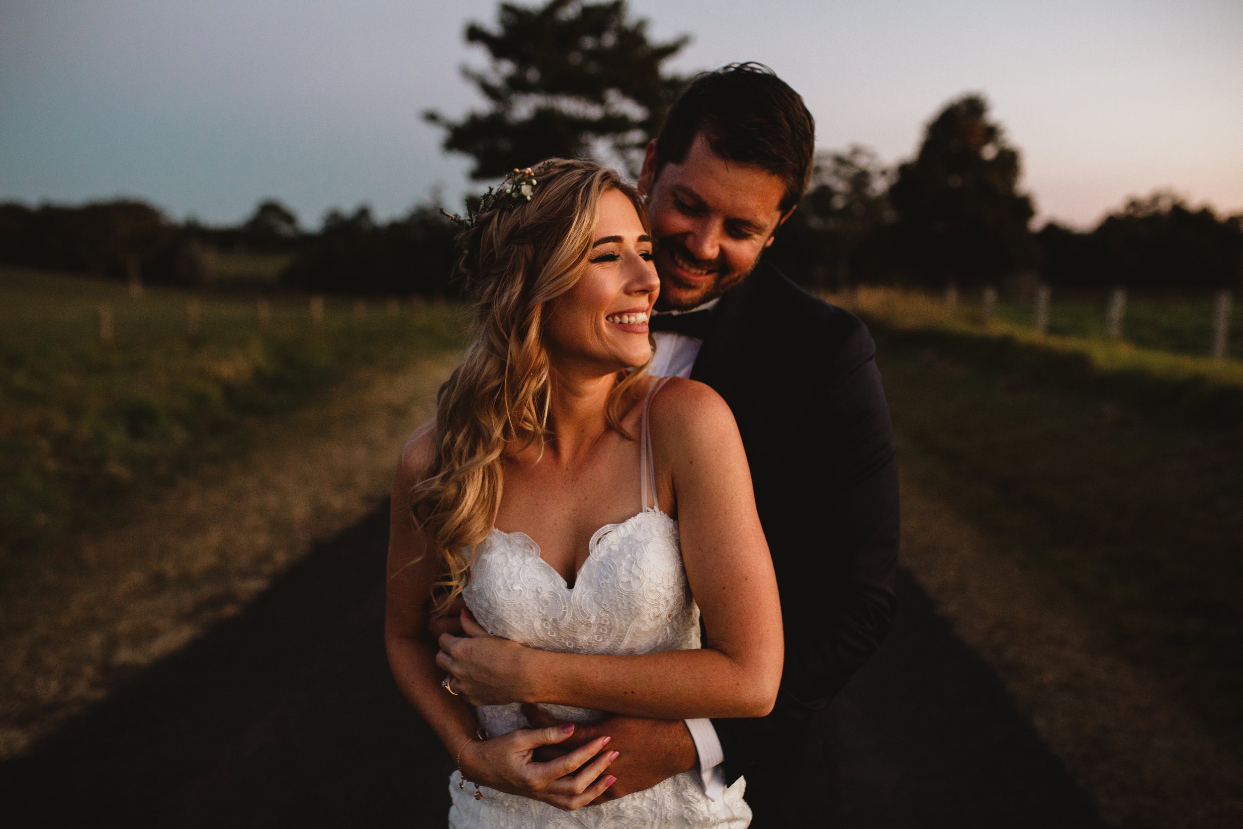 A happy newlywed couple embracing outdoors during sunset, with trees and a pathway visible in the background.