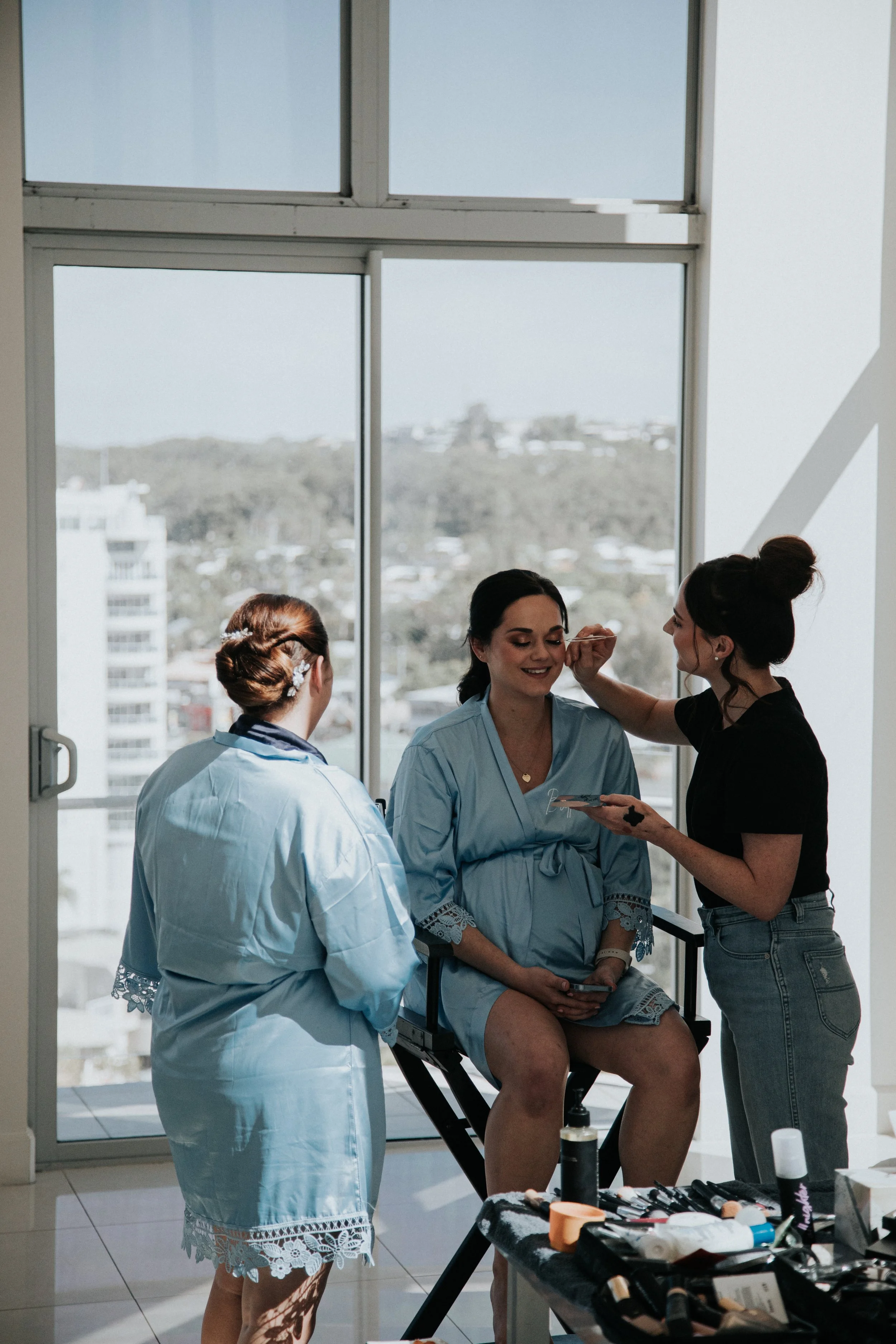 A woman in a blue robe sits on a chair while two makeup artists apply makeup to her face. They are in a bright room with large windows overlooking a cityscape.