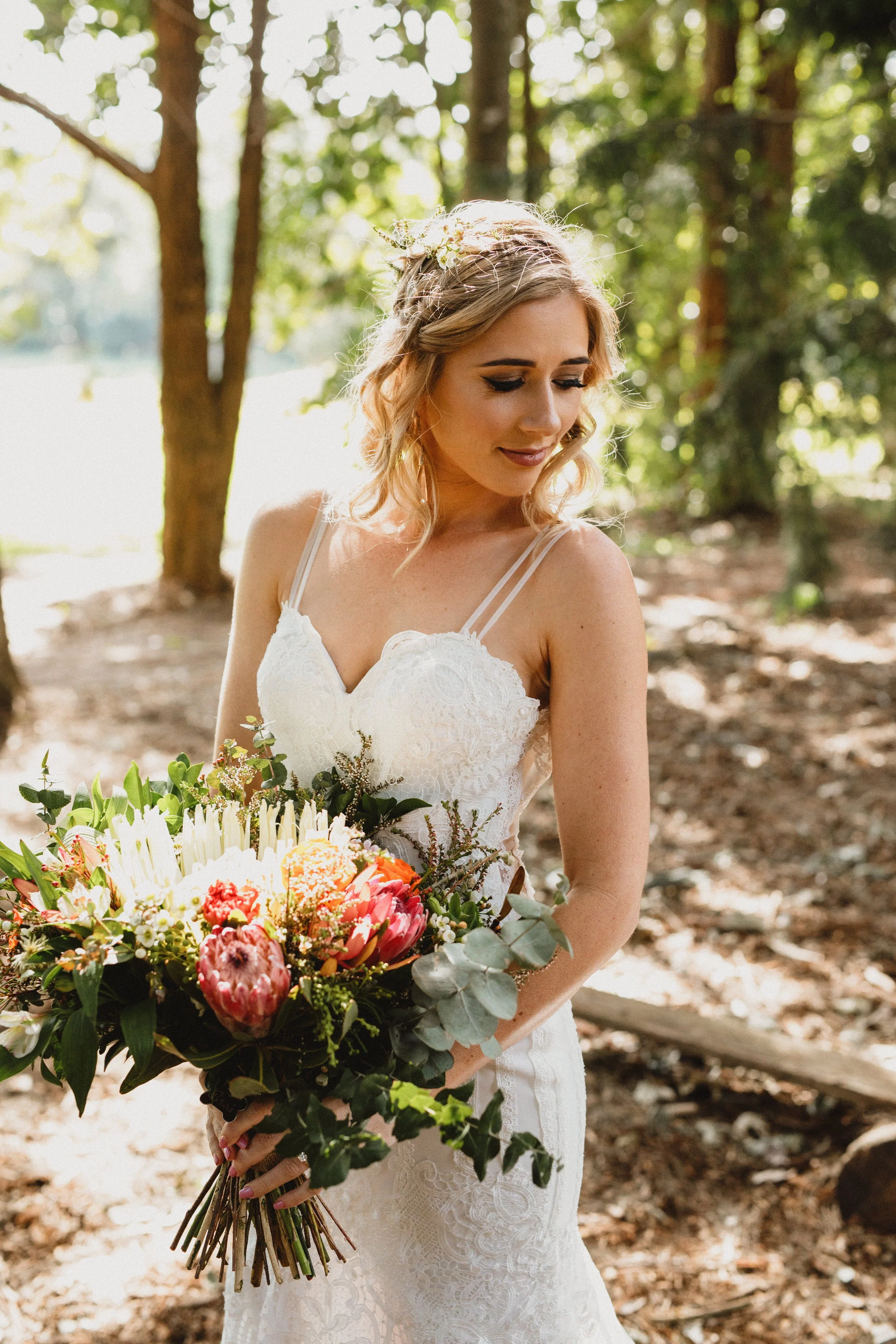 A bride in a white lace wedding dress holding a colorful bouquet of flowers, standing outdoors in a wooded area, with sunlight filtering through the trees.