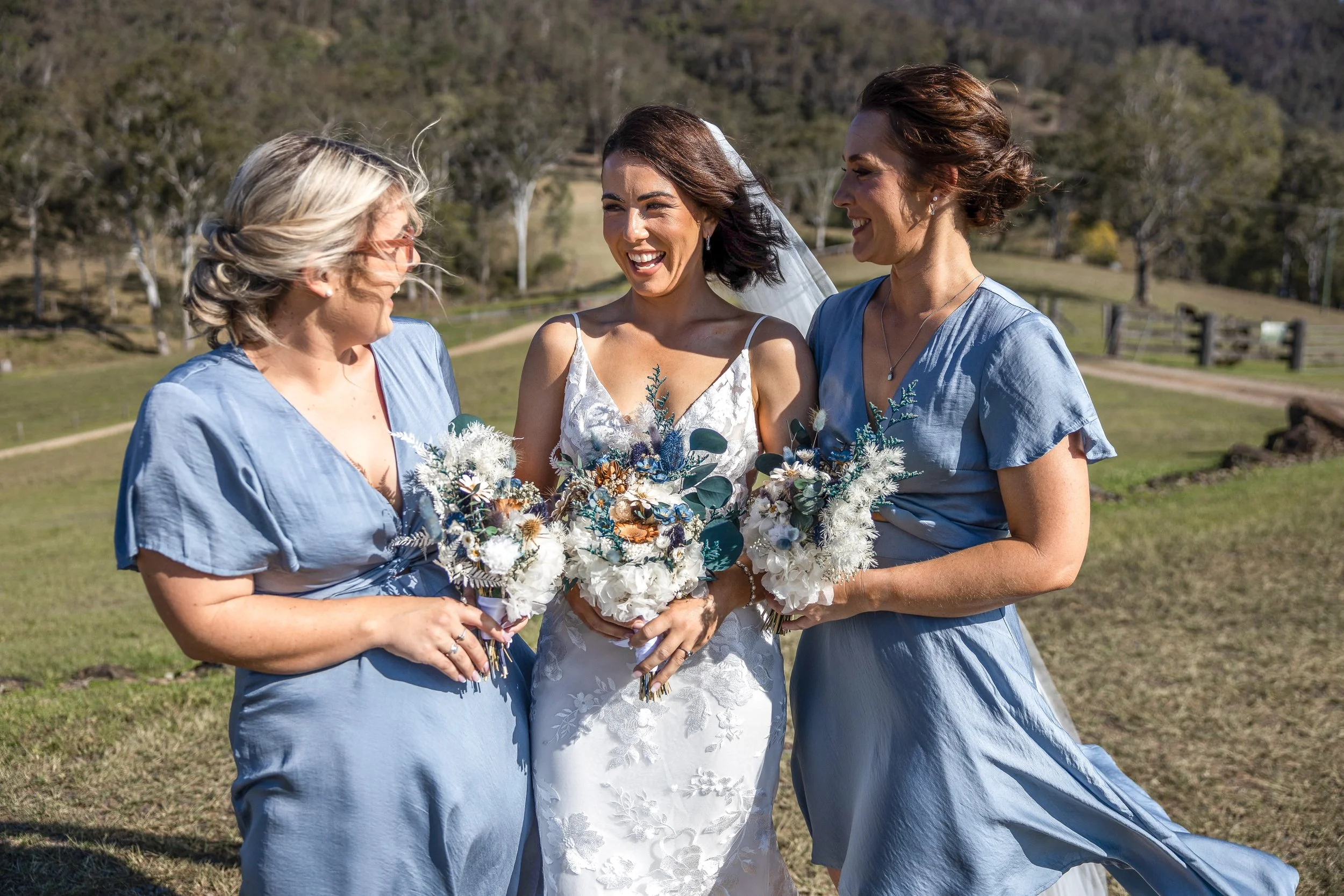 Three women at a wedding outdoors, with the bride in the center wearing a white dress and veil, and two women in matching blue dresses on each side, holding bouquets and smiling.