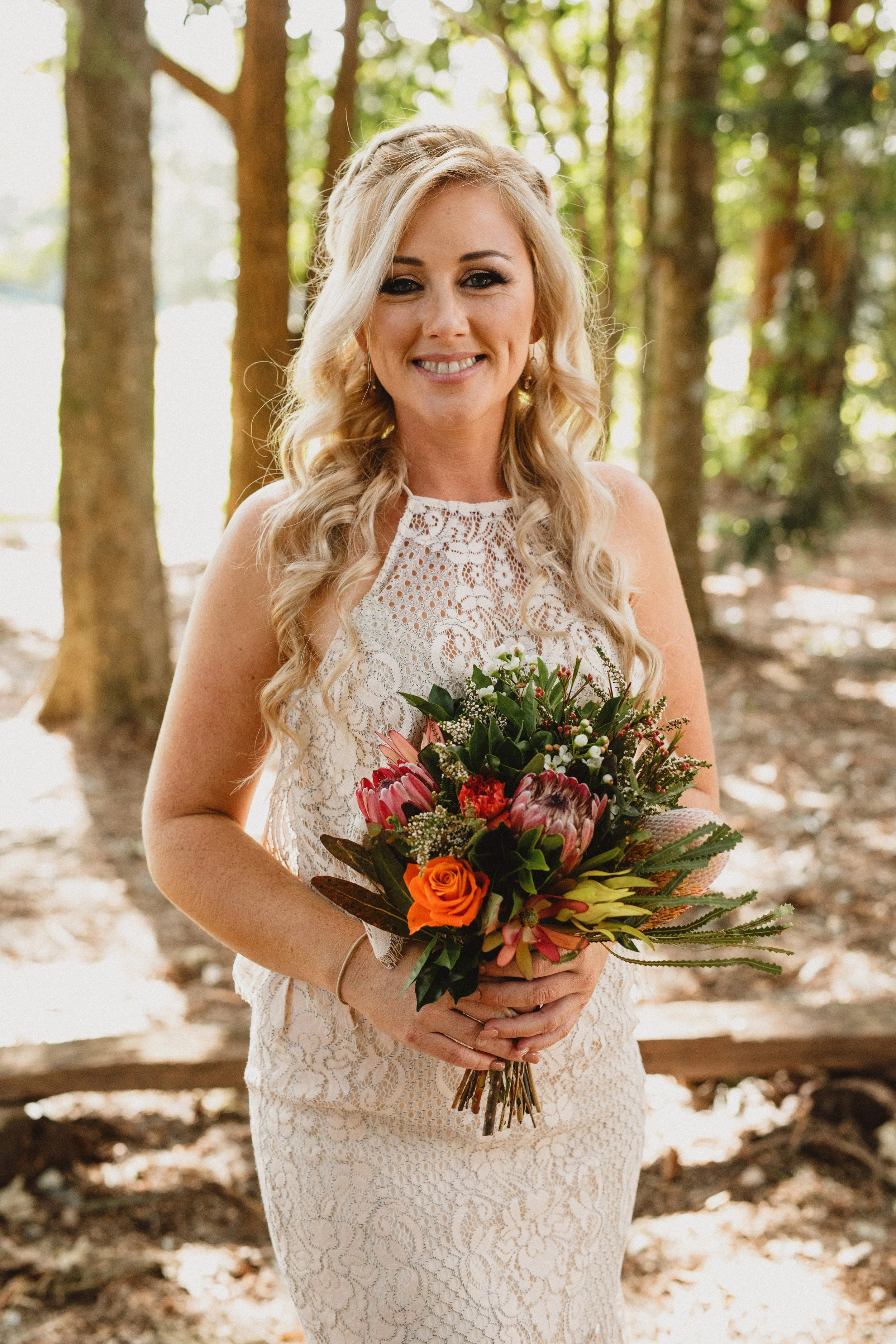 A woman in a white, lace dress holding a colorful bouquet of flowers in a forest setting.