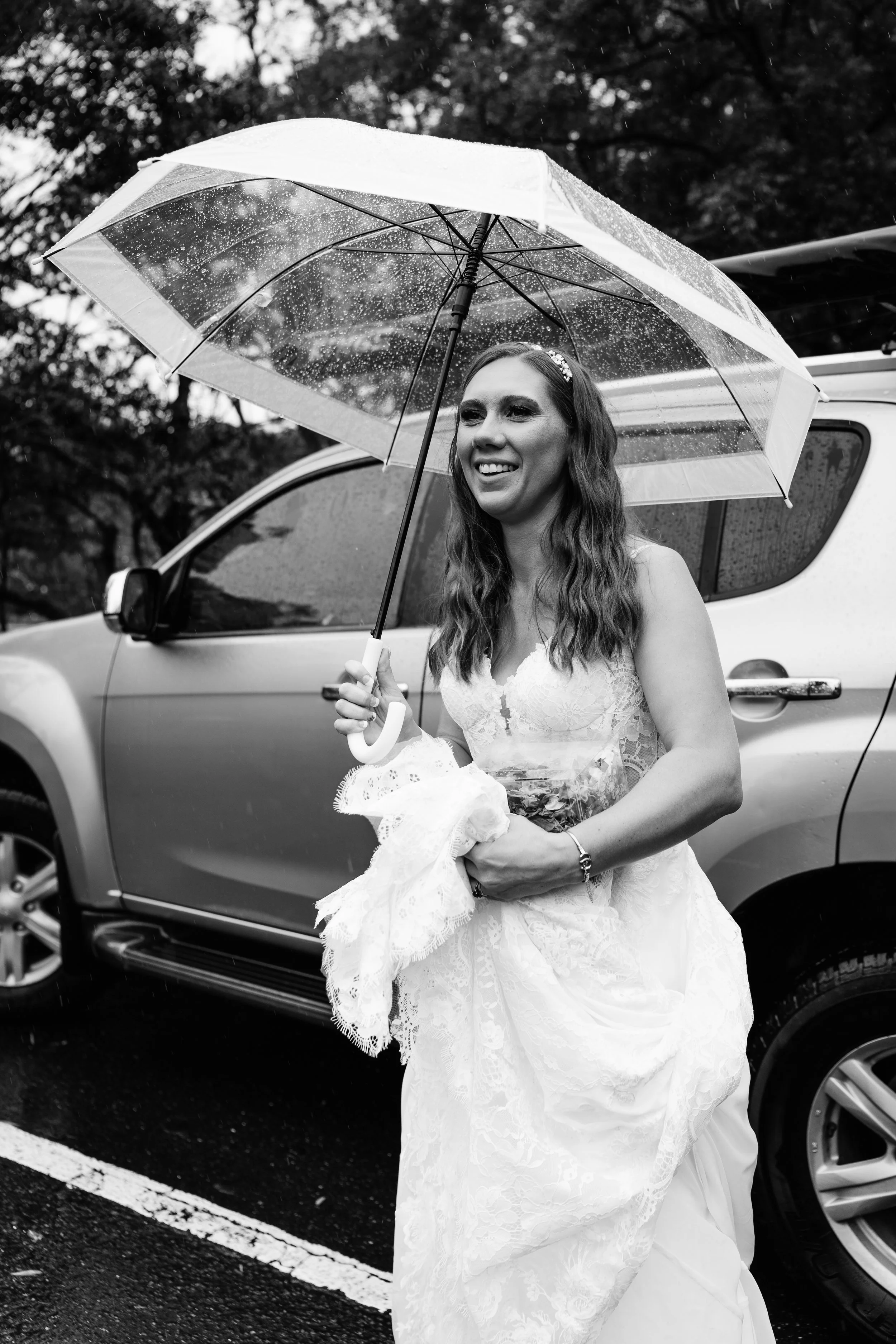 A smiling woman in a wedding dress holding a bouquet under a transparent umbrella on a rainy day, standing next to a car.