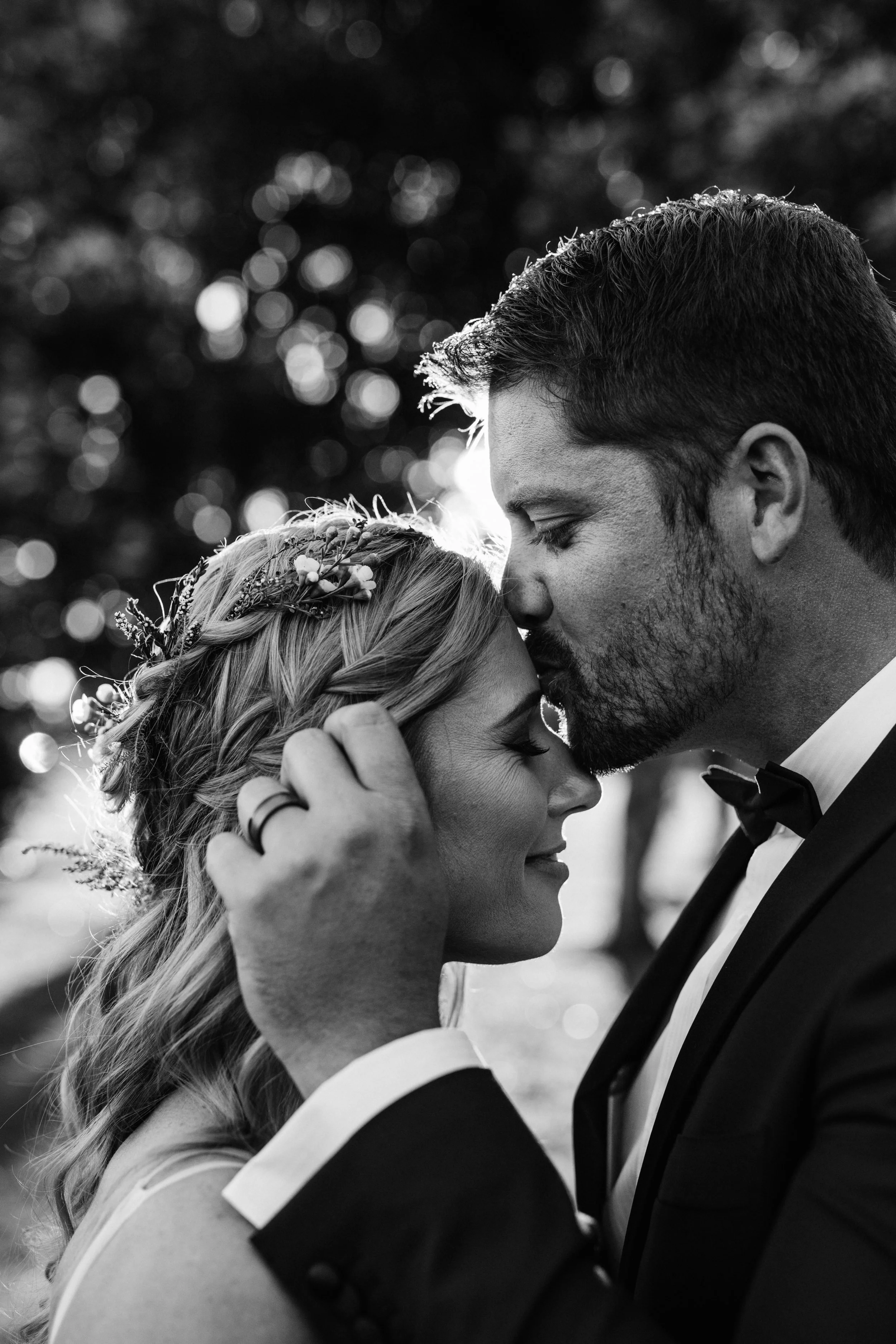 A black and white photo of a couple with their foreheads touching, eyes closed, moment of intimate connection outdoors, groom in a suit and bow tie, bride with floral headband.