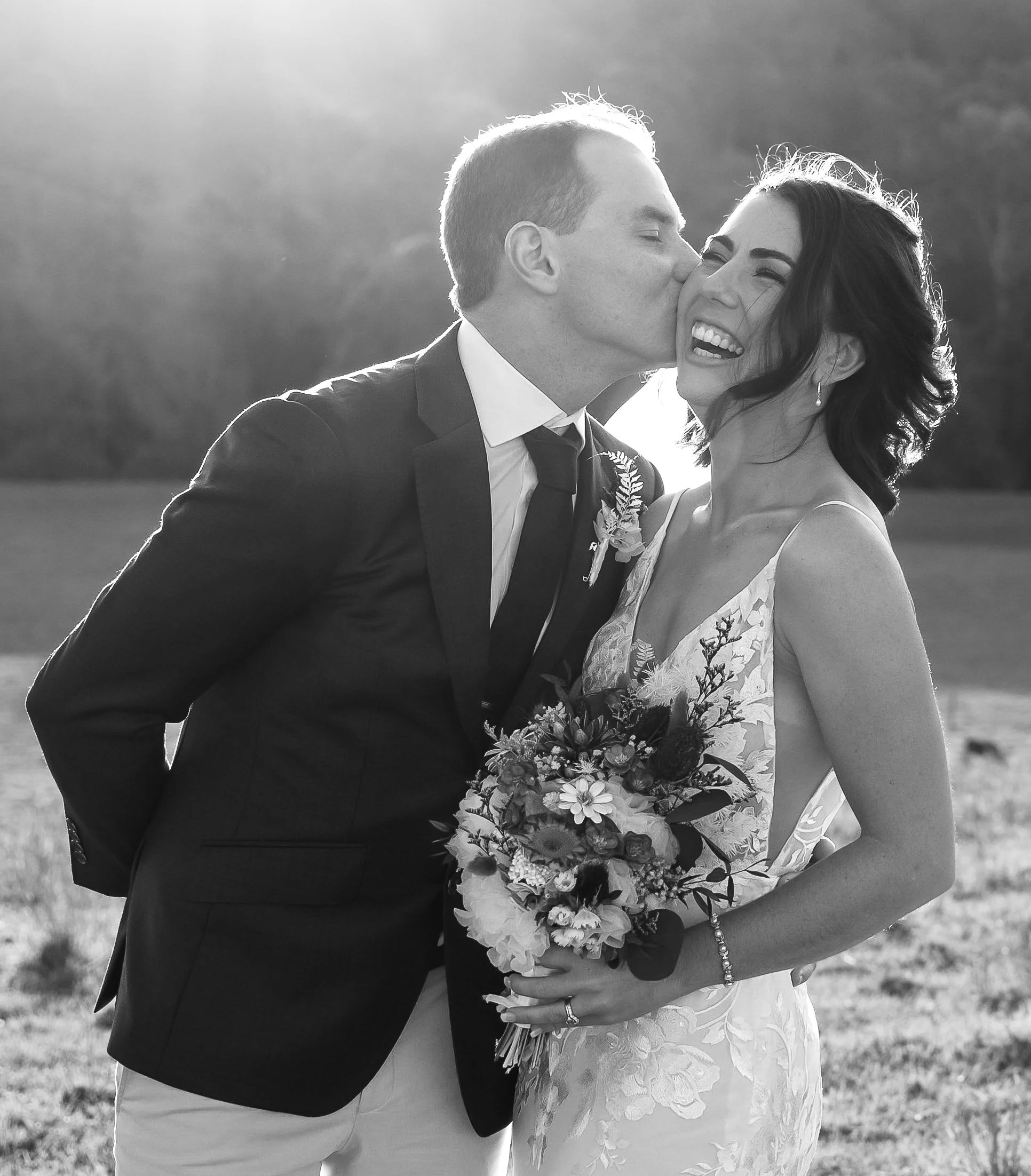 A black and white photo of a newlywed couple. The groom is kissing the bride's cheek, who is smiling and holding a bouquet of flowers outdoors in a field with trees in the background.