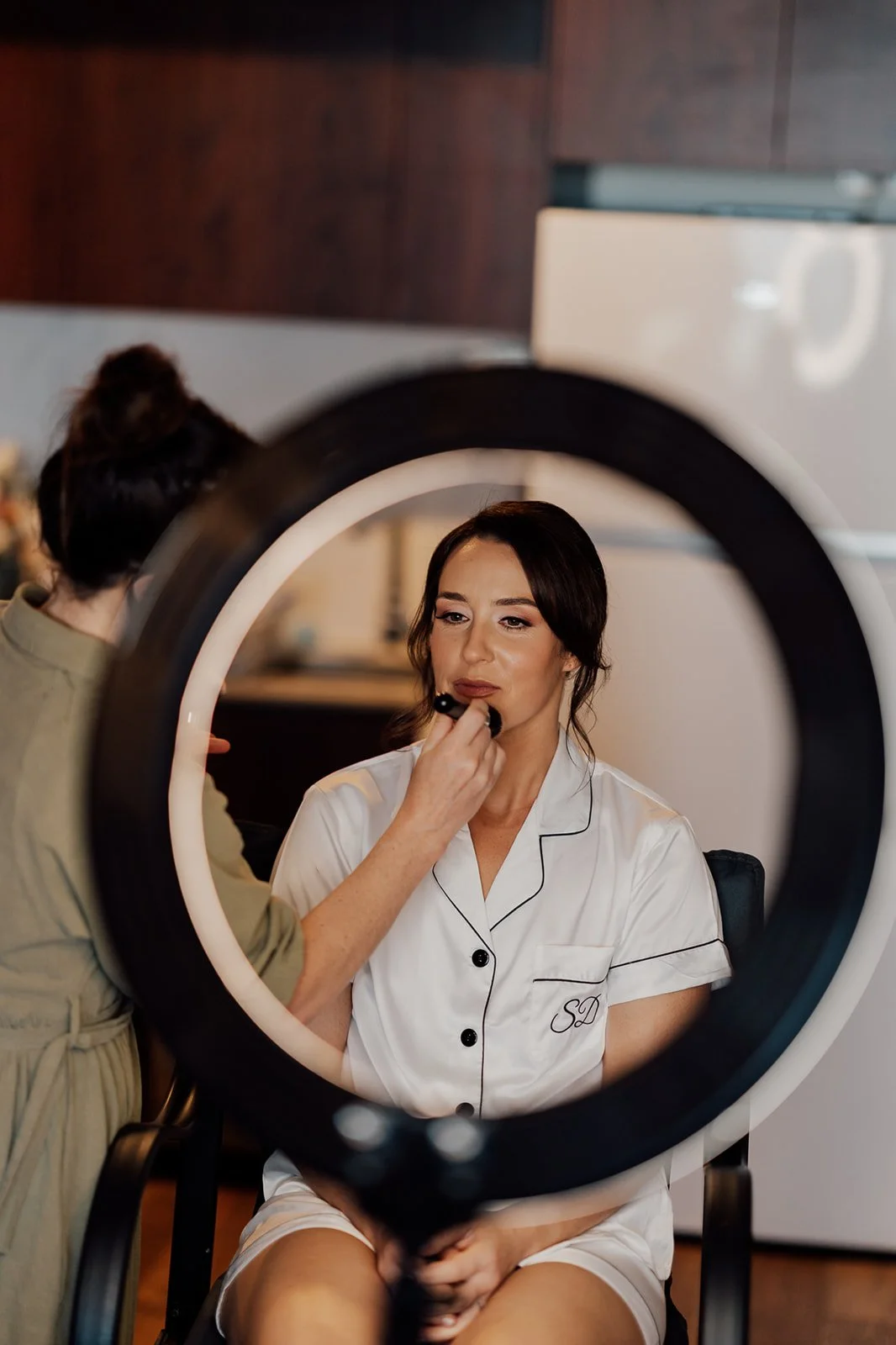 A woman is having her makeup done while sitting in a room, seen through a makeup ring light.