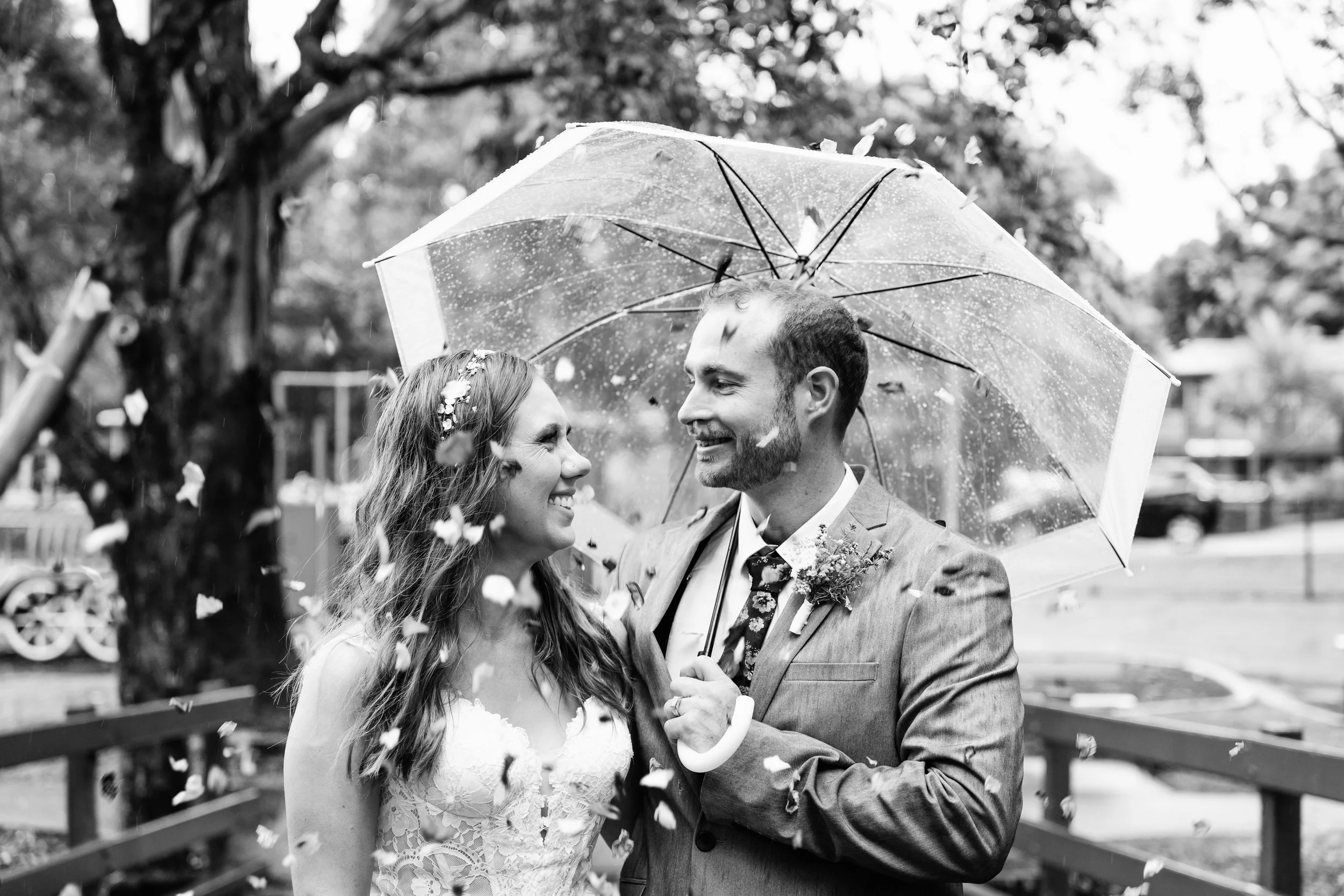 Black and white photo of a happy couple standing under an umbrella during a rainy outdoor wedding, with confetti falling around them.