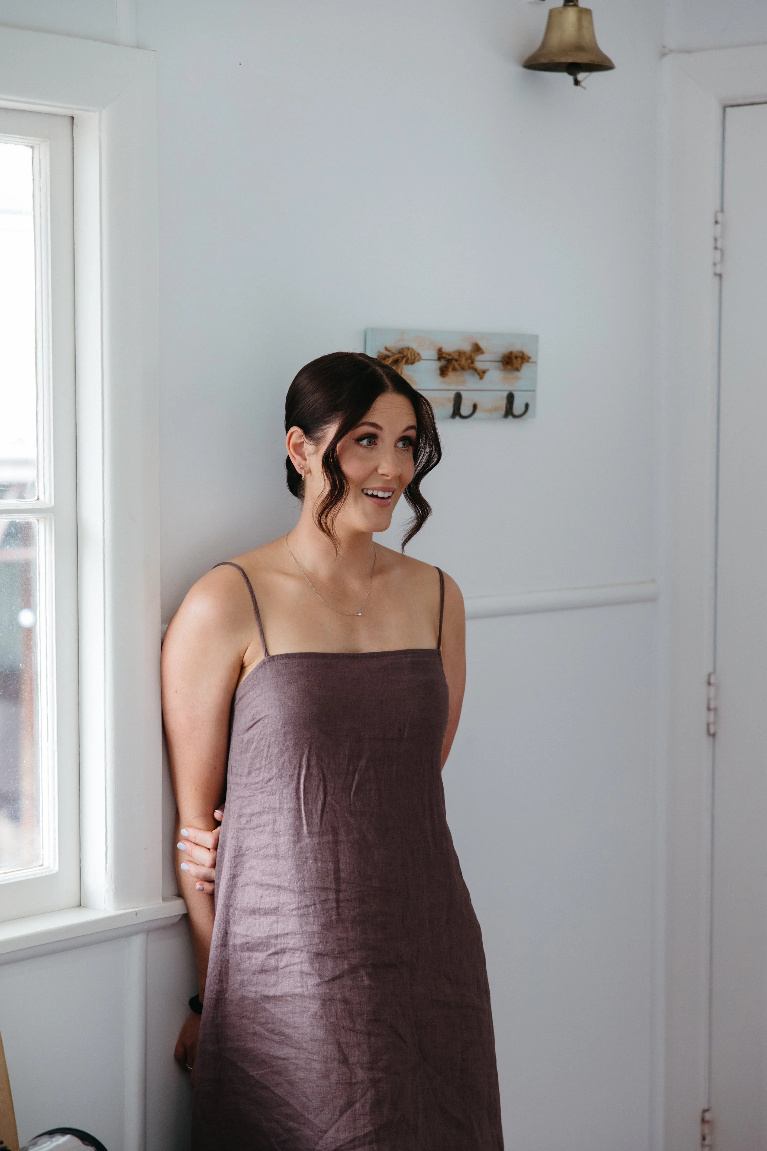 A woman with dark brown wavy hair in a low bun, wearing a sleeveless brown dress, standing by a window inside a white-walled room, smiling and looking to the side.
