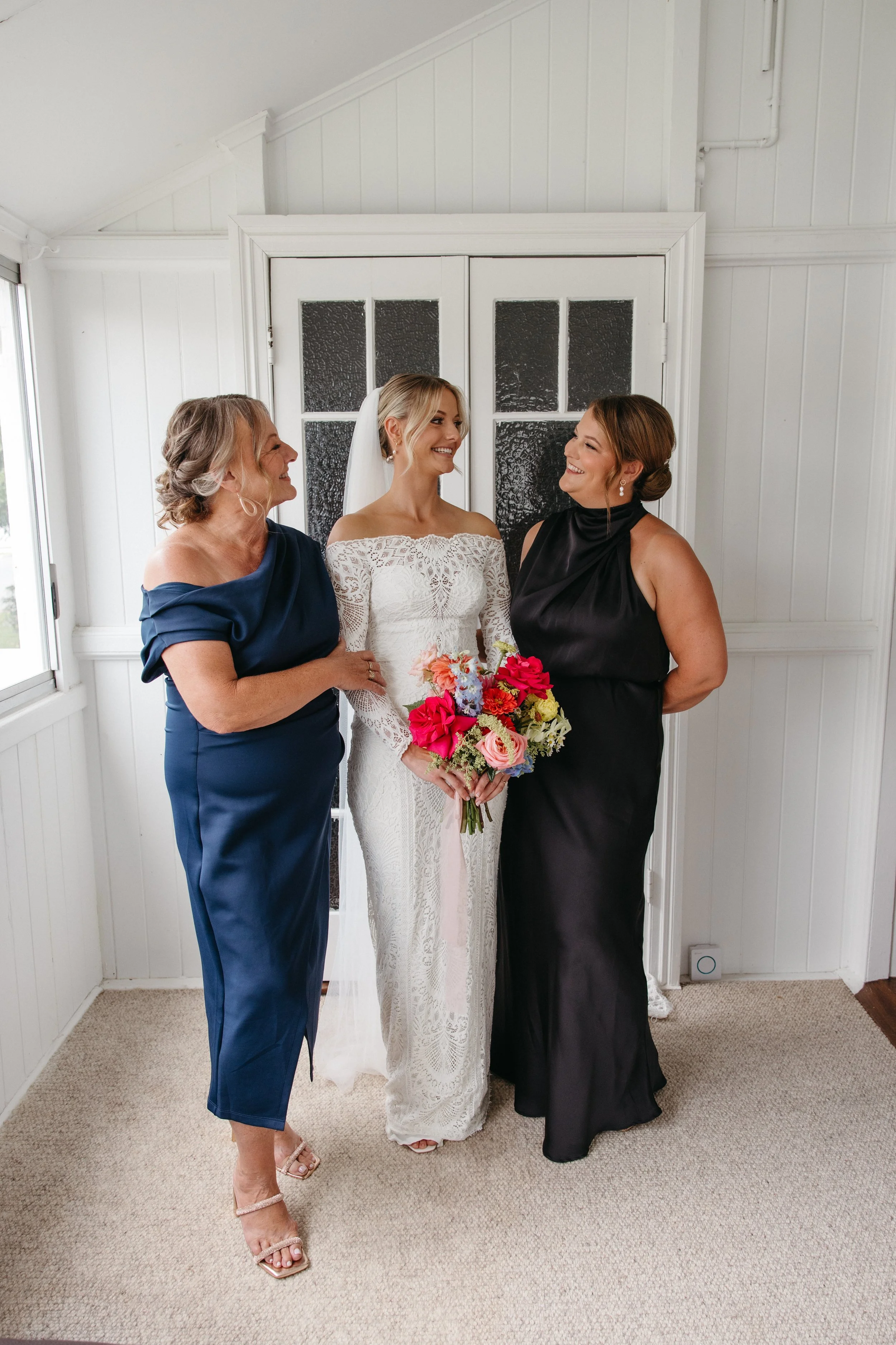 Bride with blonde hair wearing a lace wedding dress, holding a colorful bouquet, standing between two women in elegant dresses inside a white room.