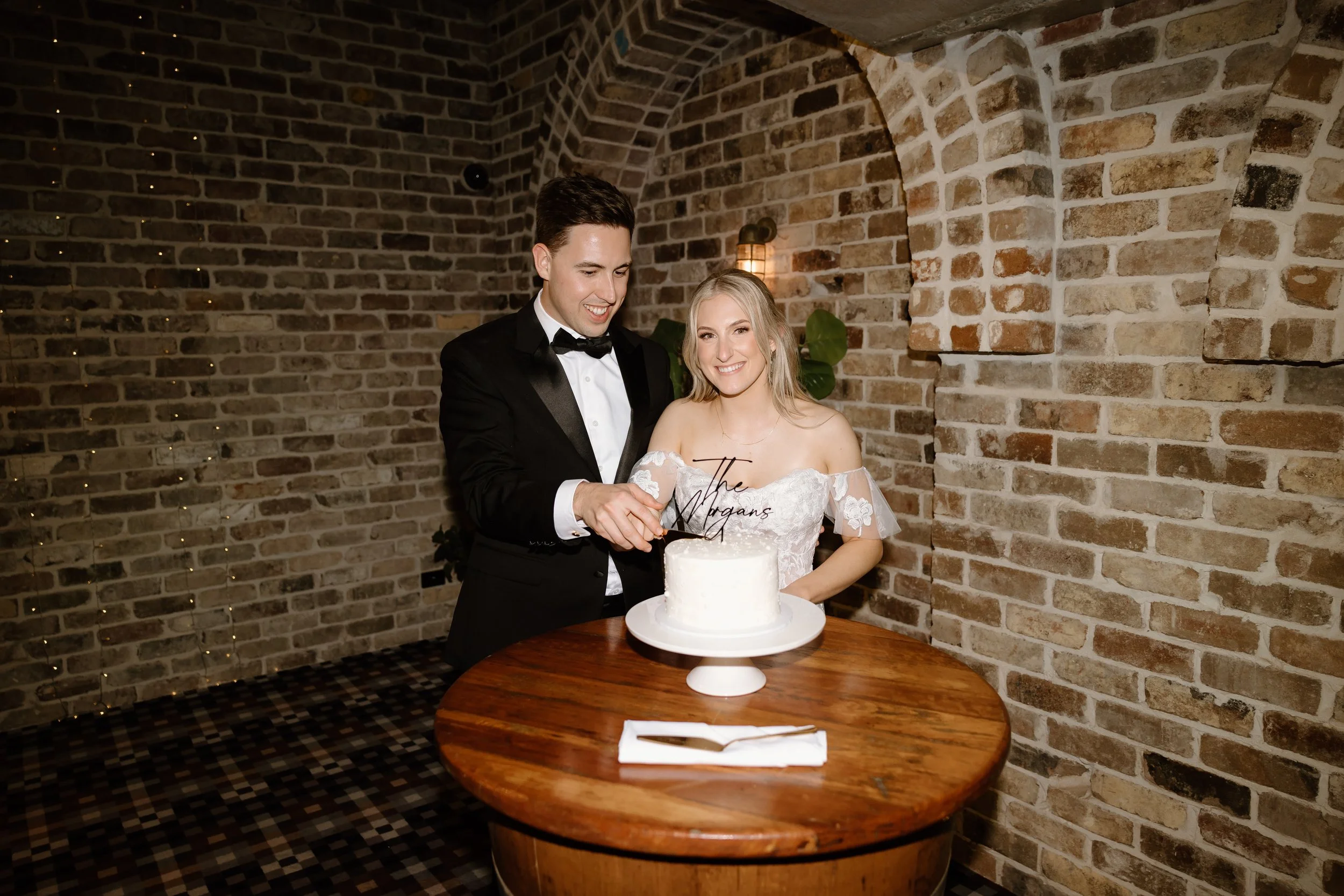 A newlywed couple in formal attire cutting a white frosted wedding cake with a topper that says 'The Morgans' in a rustic brick-walled setting.